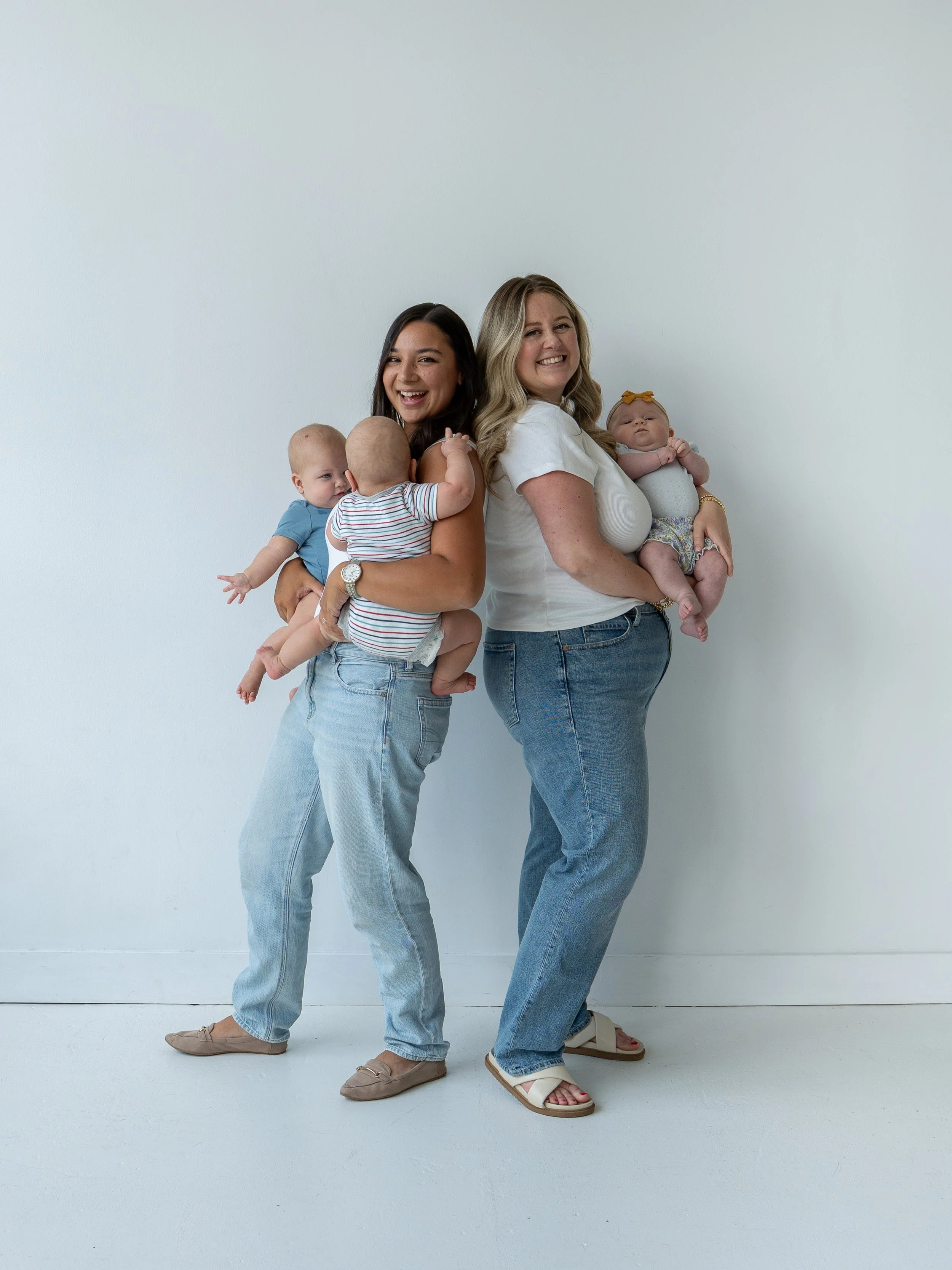 Two women holding three babies, standing against a white wall, smiling and showing affection.