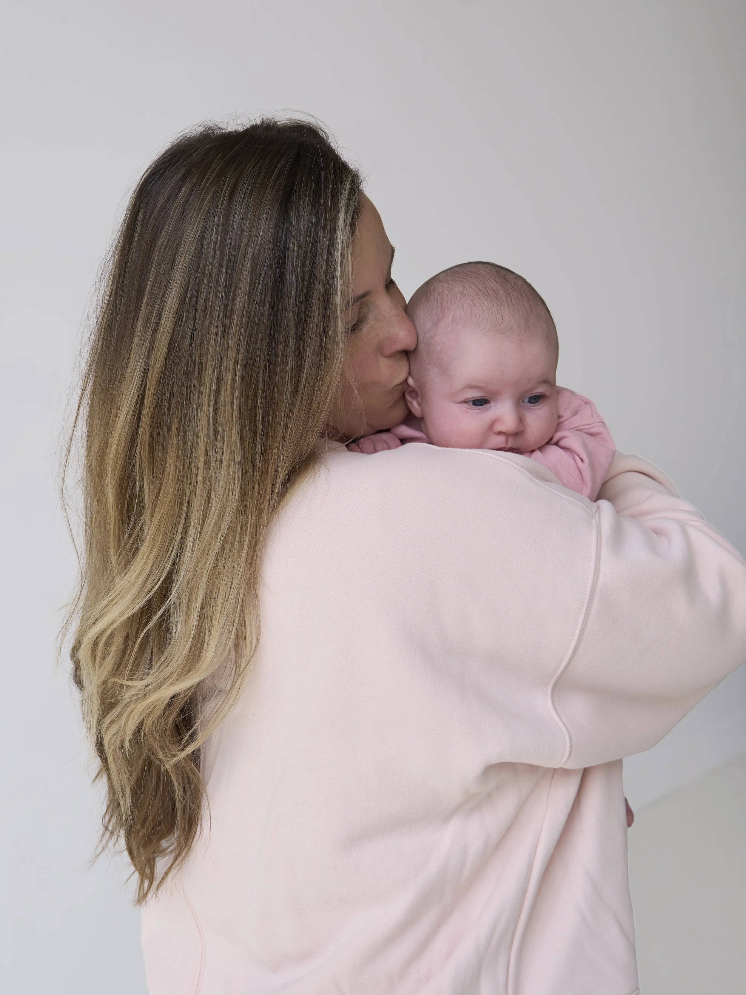A woman with long, wavy, light brown hair holding a baby with a bald head and blue eyes, both with their eyes closed, against a plain light-colored background.