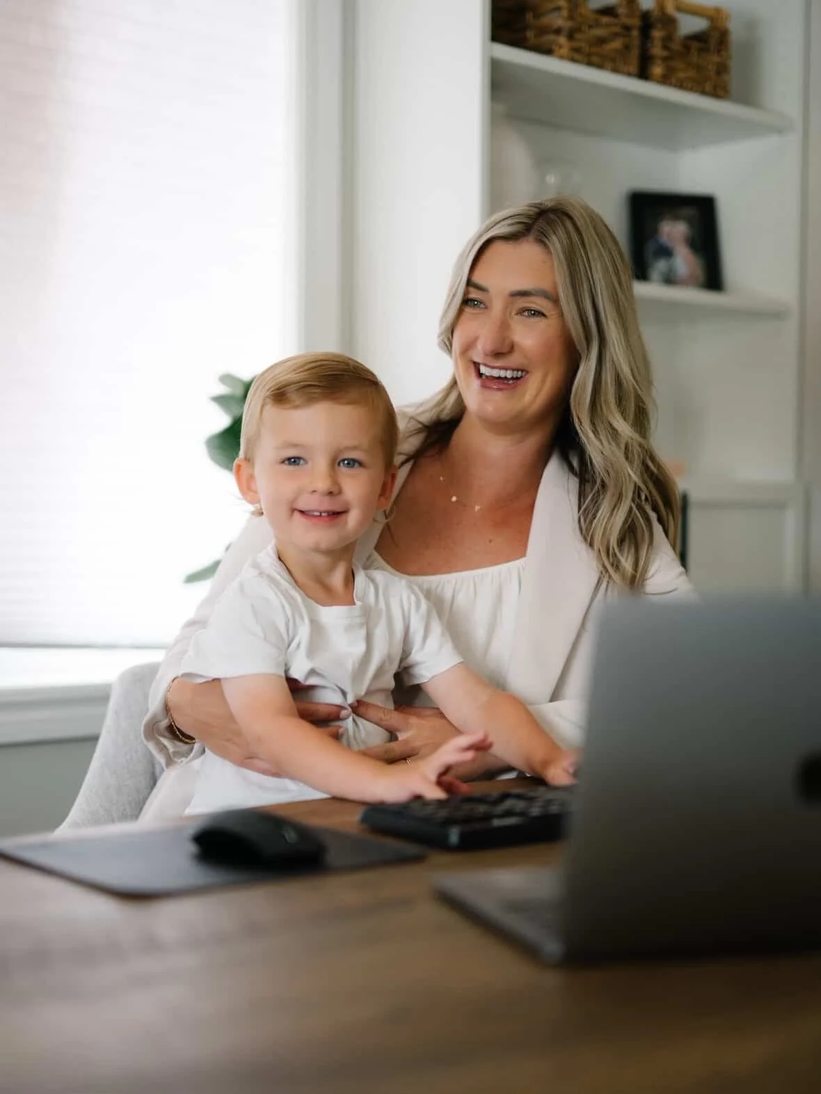 A woman and a young boy sitting at a desk, smiling at a laptop computer in a home setting.