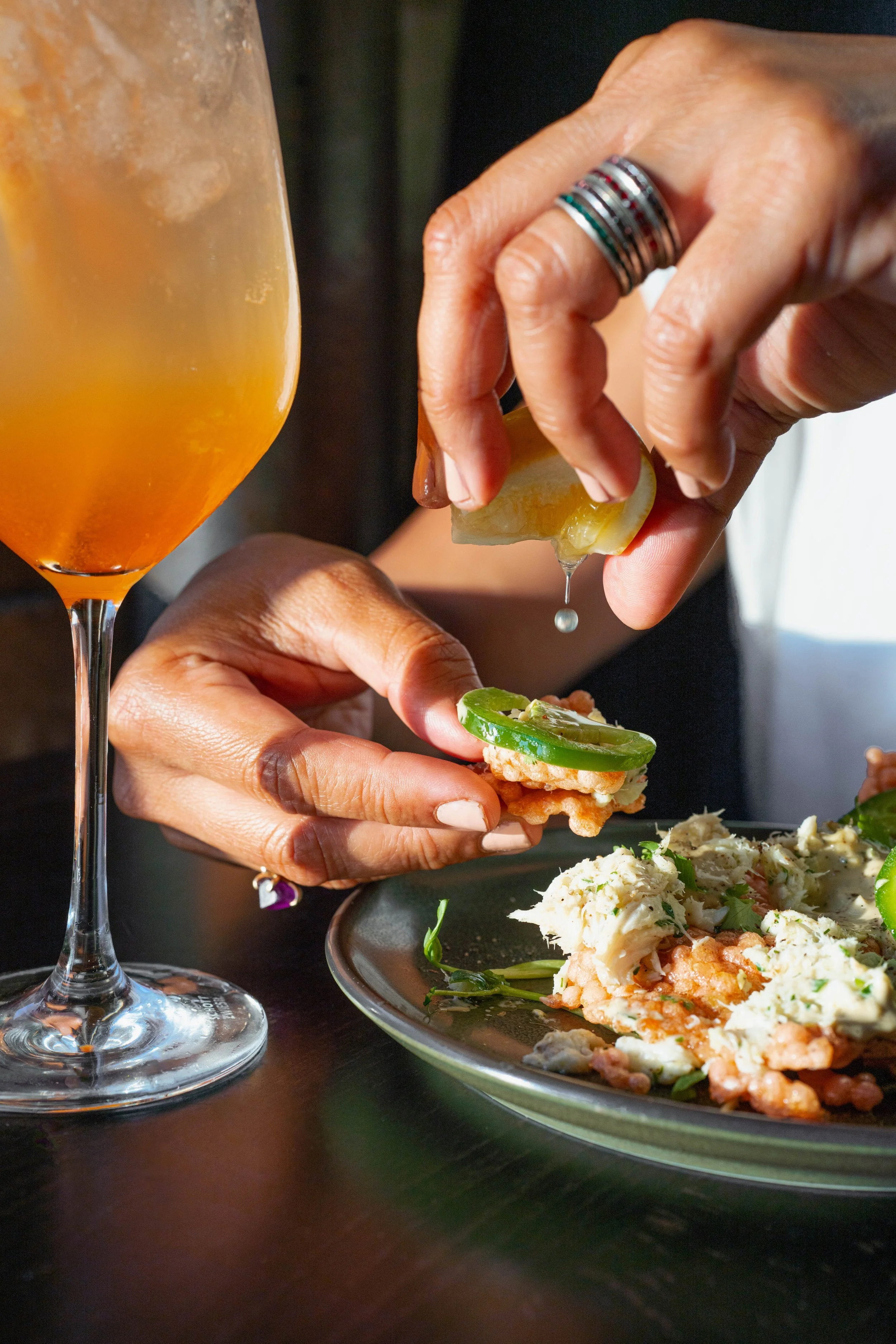 A person squeezing a lemon over a crisp topped with seafood and jalapeño on a black plate, with an orange cocktail in a glass nearby.