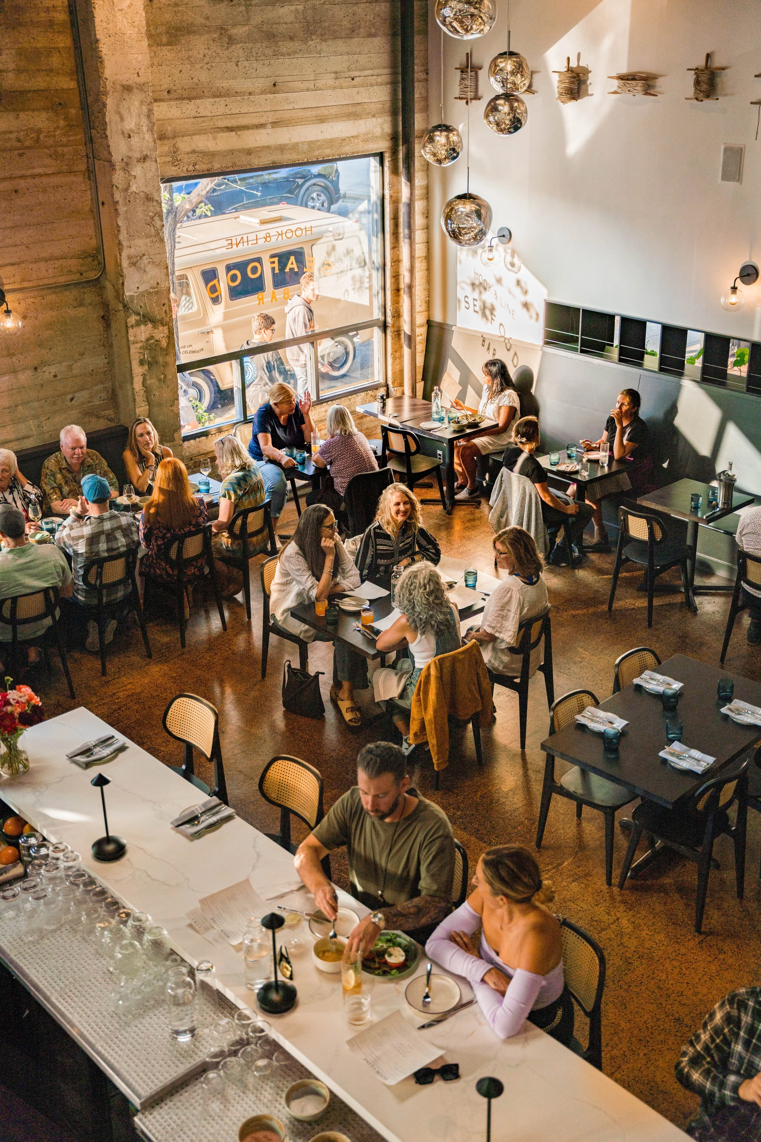 Overhead view of a busy restaurant with people dining at various tables, modern decor, and large windows with sunlight streaming in.
