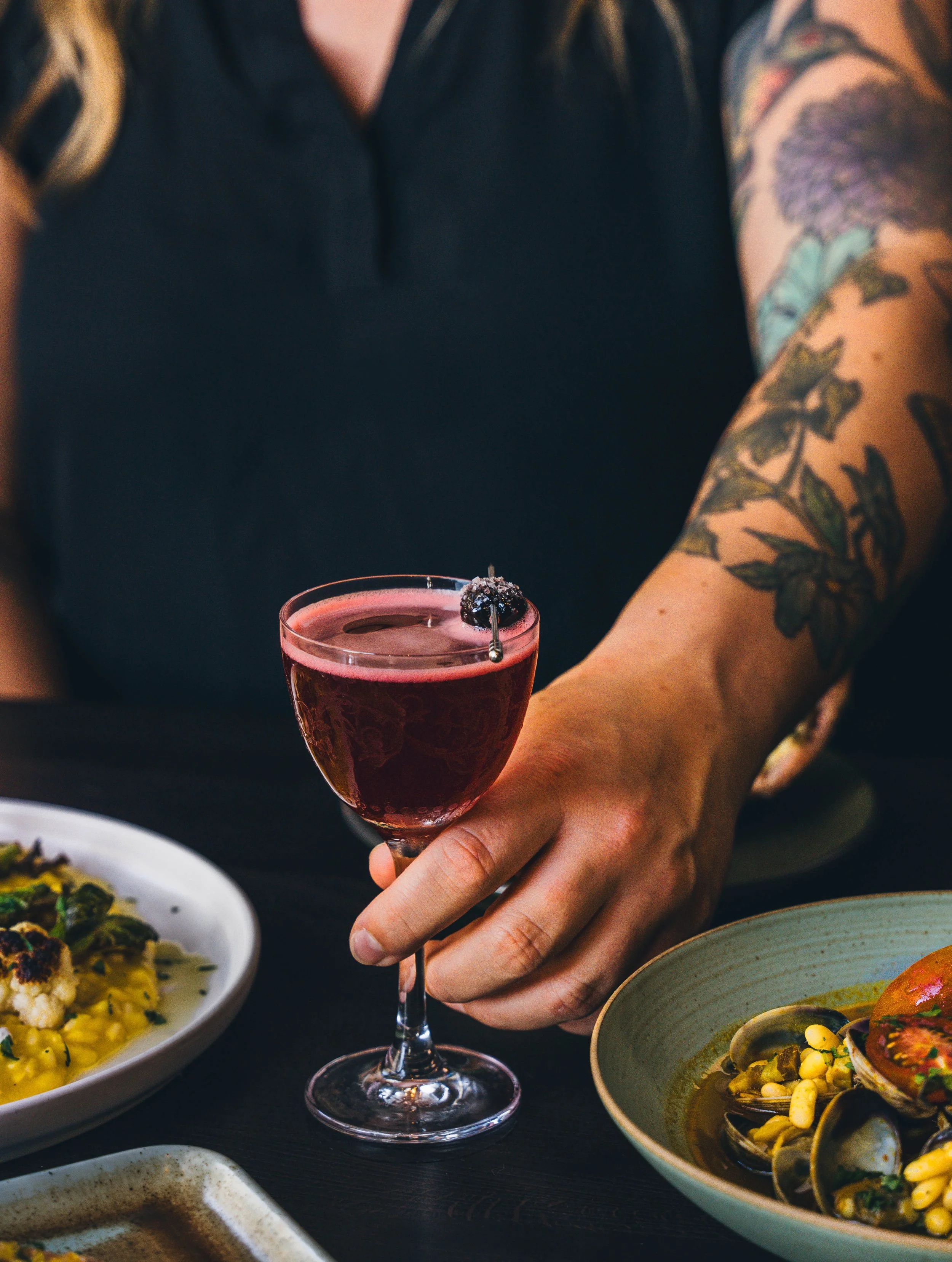 Tattooed person holding a pink cocktail in a stemmed glass, with dishes of food on the table.