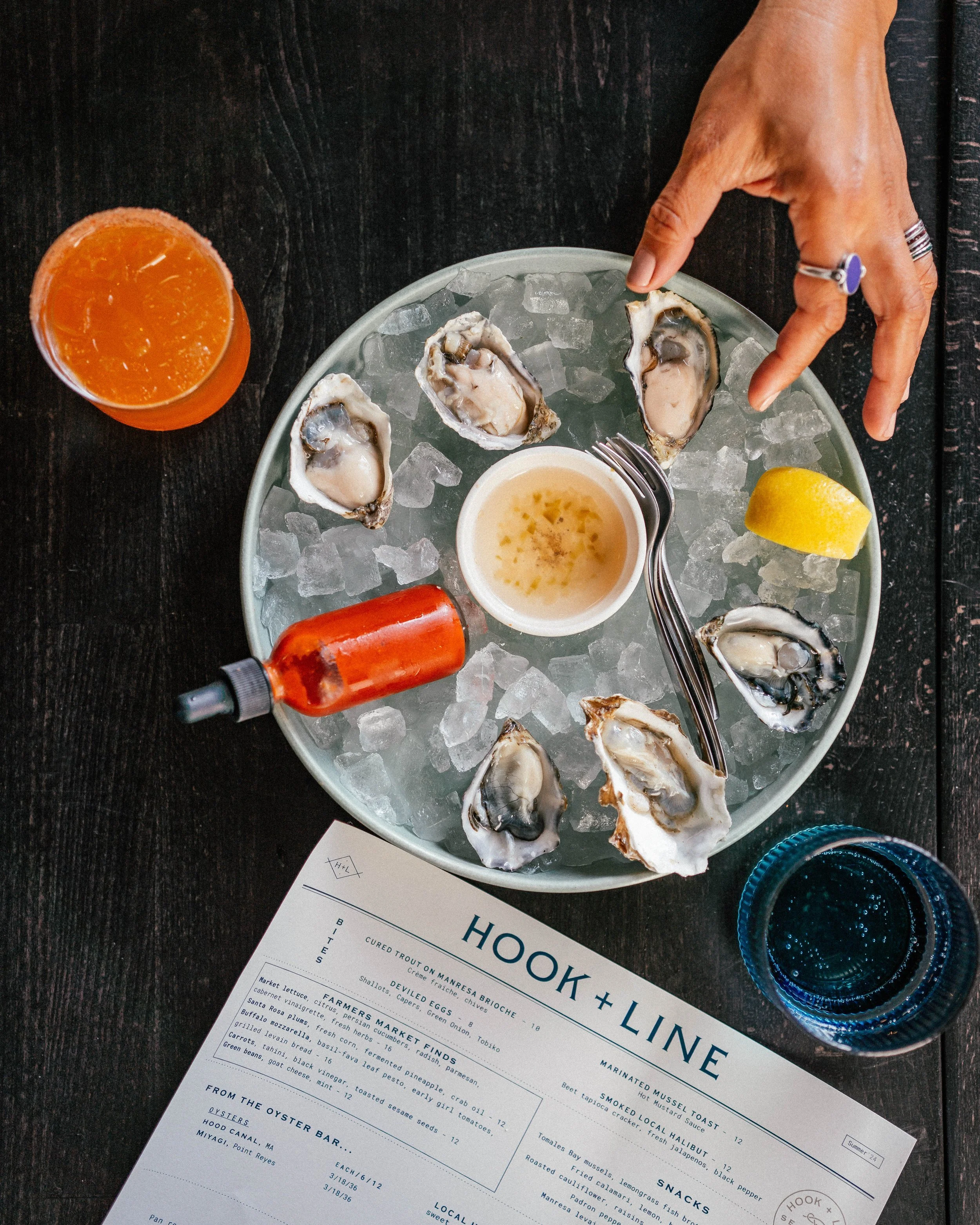 A plate of oysters on ice with lemon wedge, red sauce bottle, and a menu titled 'Hook + Line' on a dark table. A person’s hand reaches for an oyster. Nearby are a glass of orange beverage and a blue glass.