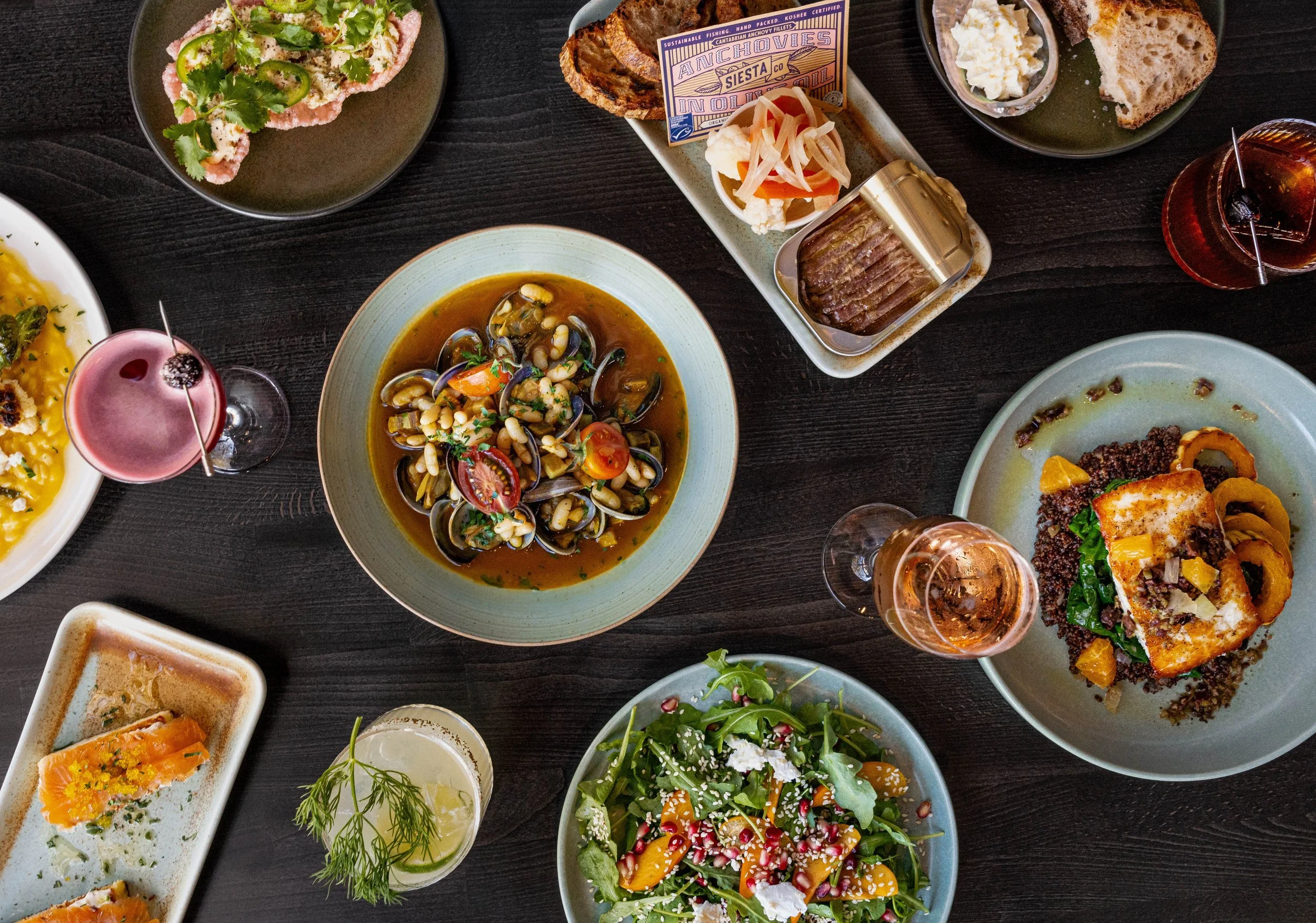 Assorted dishes on a dark table including seafood stew, mixed salad, bread with spread, canned anchovies, and drinks.