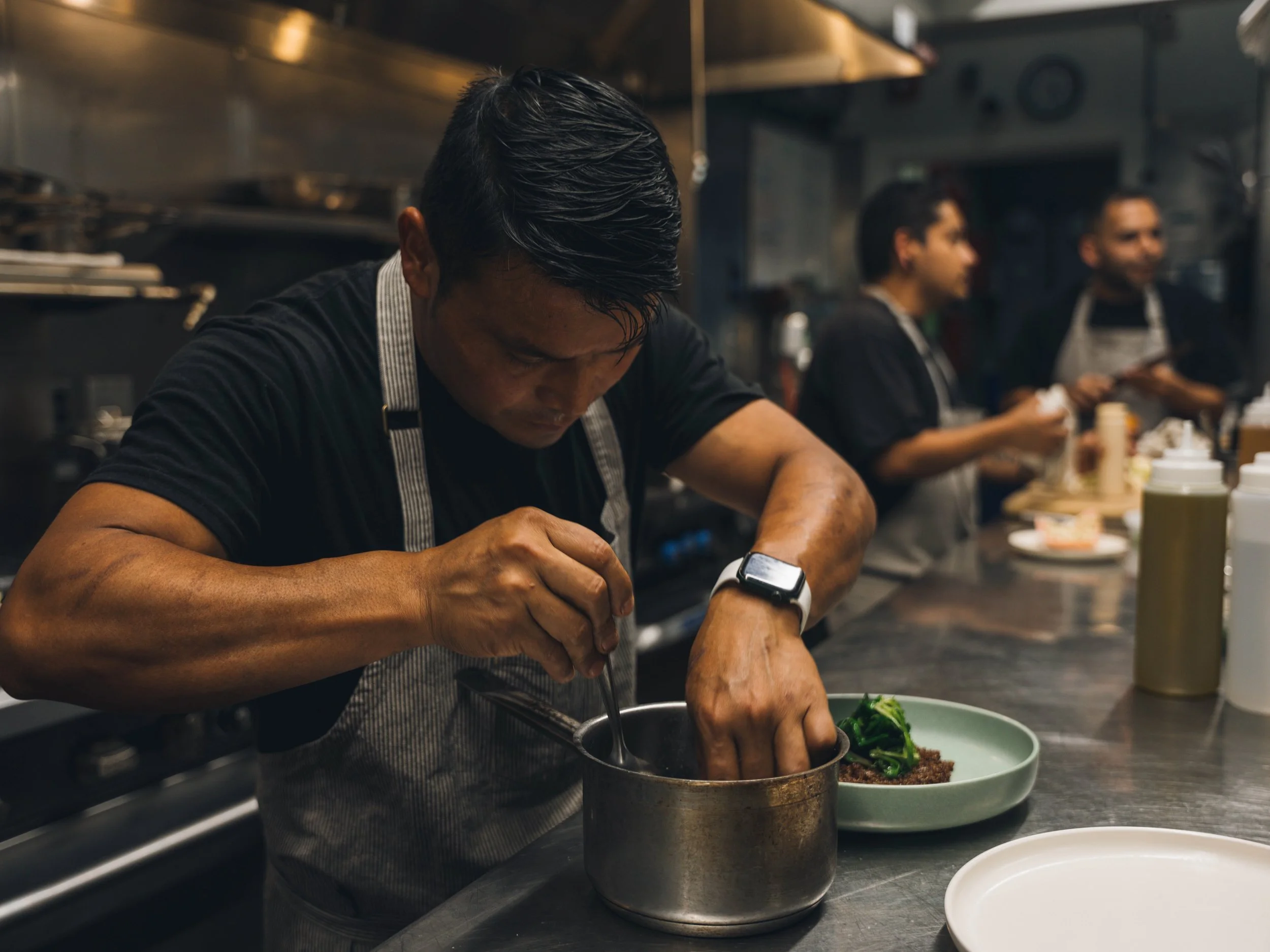 A chef in a black shirt and apron stirs a pot on a kitchen counter, while two other chefs work in the background. The kitchen has a stainless steel countertop and various utensils.