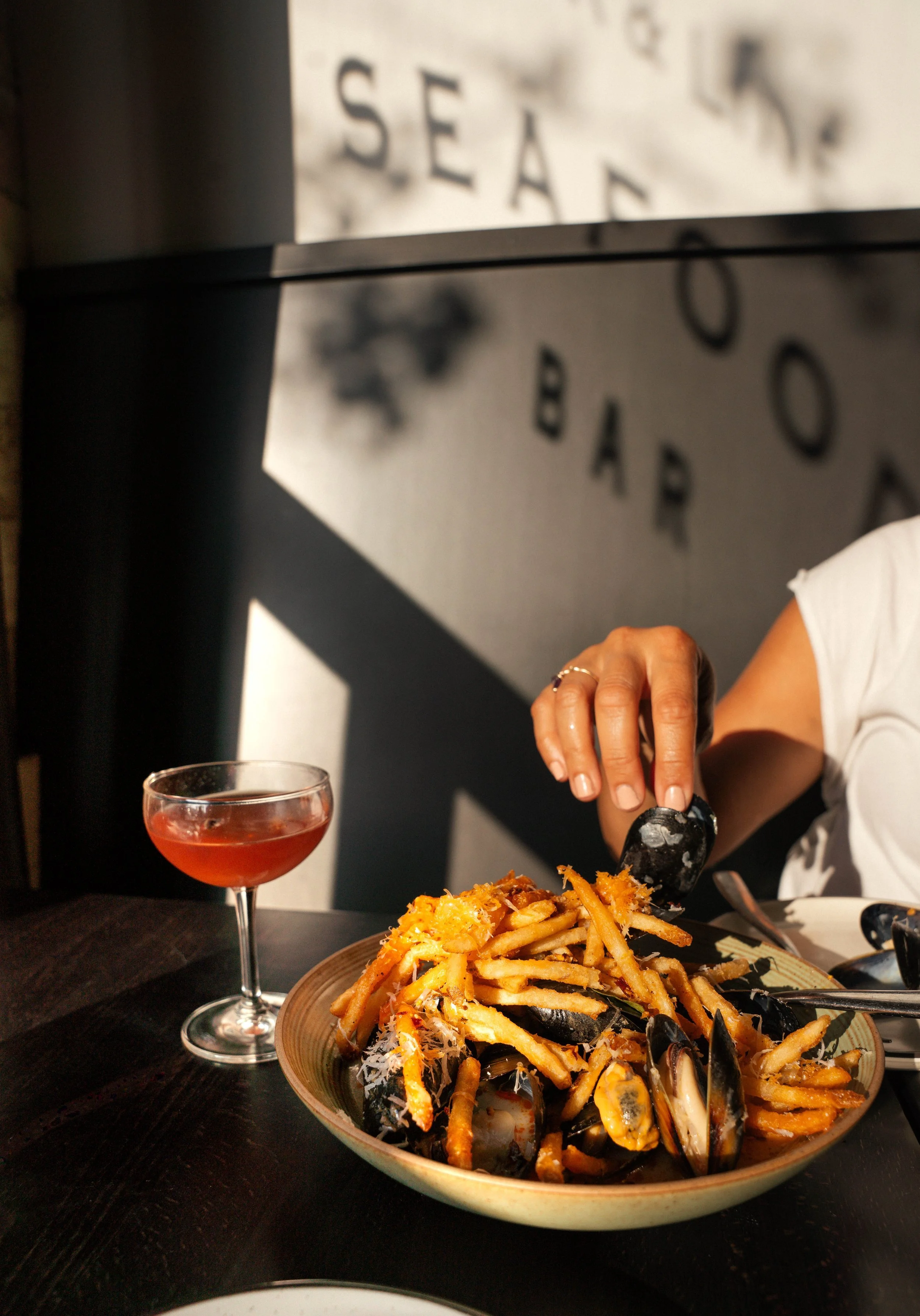 A person reaching for food in a bowl of French fries topped with mussels, next to a cocktail in a glass at a seafood-themed restaurant.