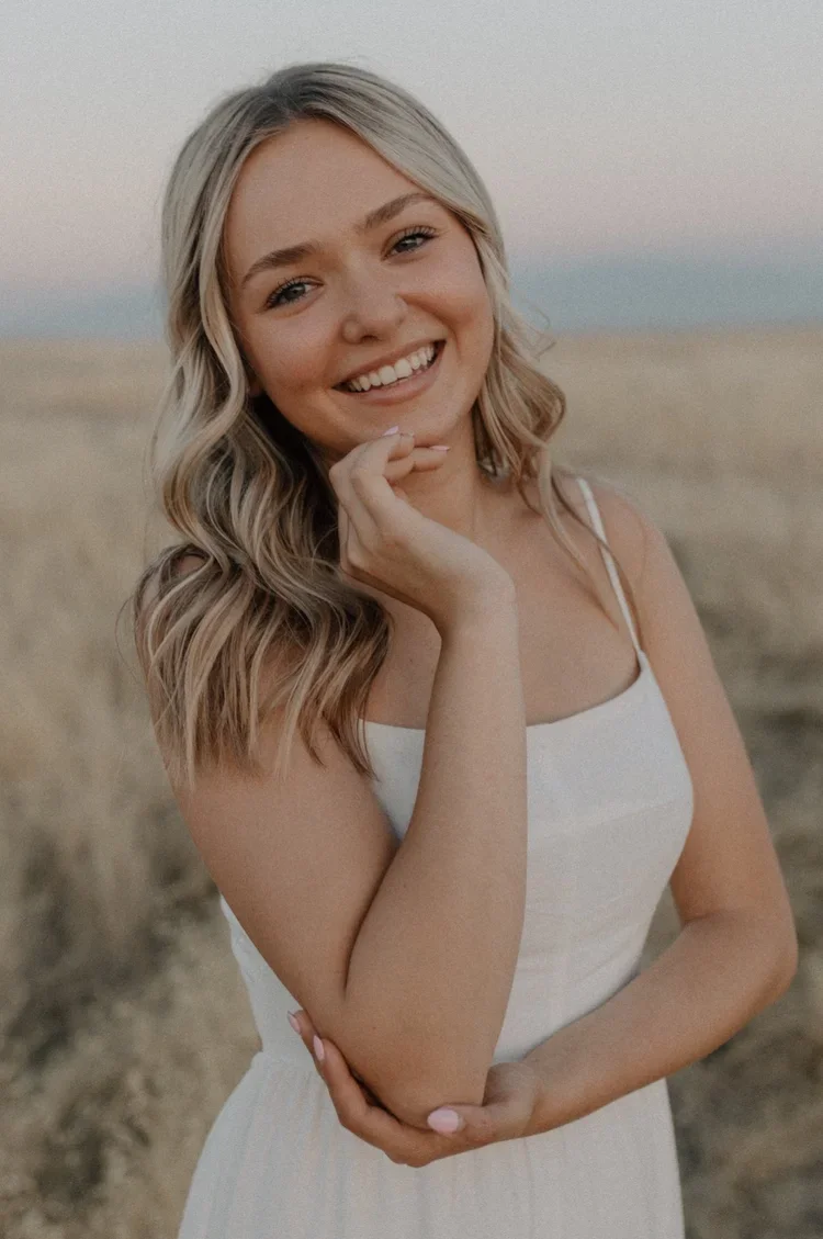 Caitlin from Wildflower Counseling Collective smiling outdoors in a natural field setting, wearing a white dress during a warm sunset portrait session.