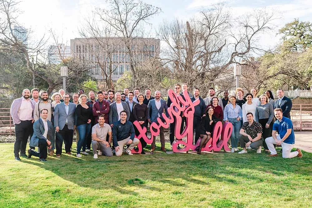 A group of people standing on a grassy area outdoors holding a large pink sign that says "You're Amazing" with trees and a building in the background.