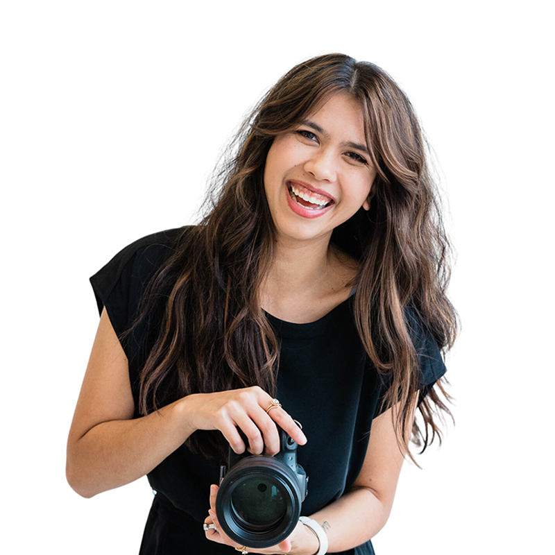 Woman with long wavy brown hair smiling, holding a camera, wearing a black shirt, standing against a white background.