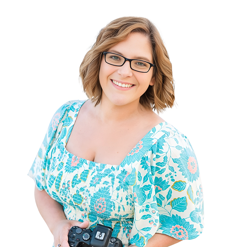 A woman with shoulder-length wavy hair, wearing black glasses and a floral patterned dress, smiling and holding a camera.
