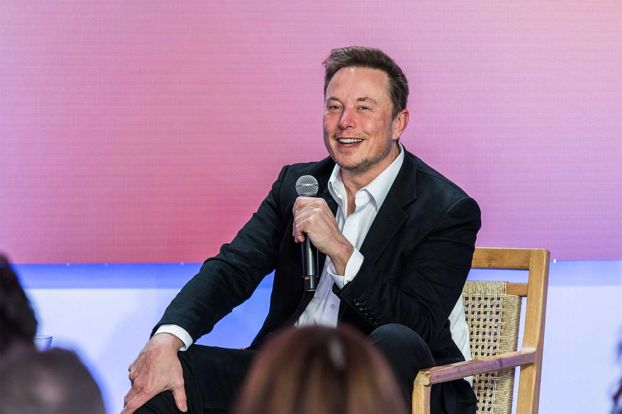 A smiling man in a black blazer and white shirt sitting on a wooden chair with a woven seat, holding a microphone, during a panel discussion.  San Francisco Bay Area corporate event and headshot photographers and videographers in Palo Alto, San Franci