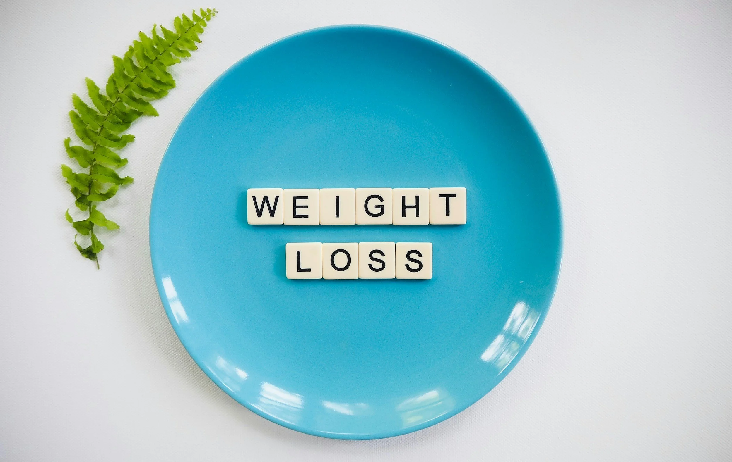 Blue plate with tiles spelling 'weight loss', accompanied by a fern leaf on a white background.