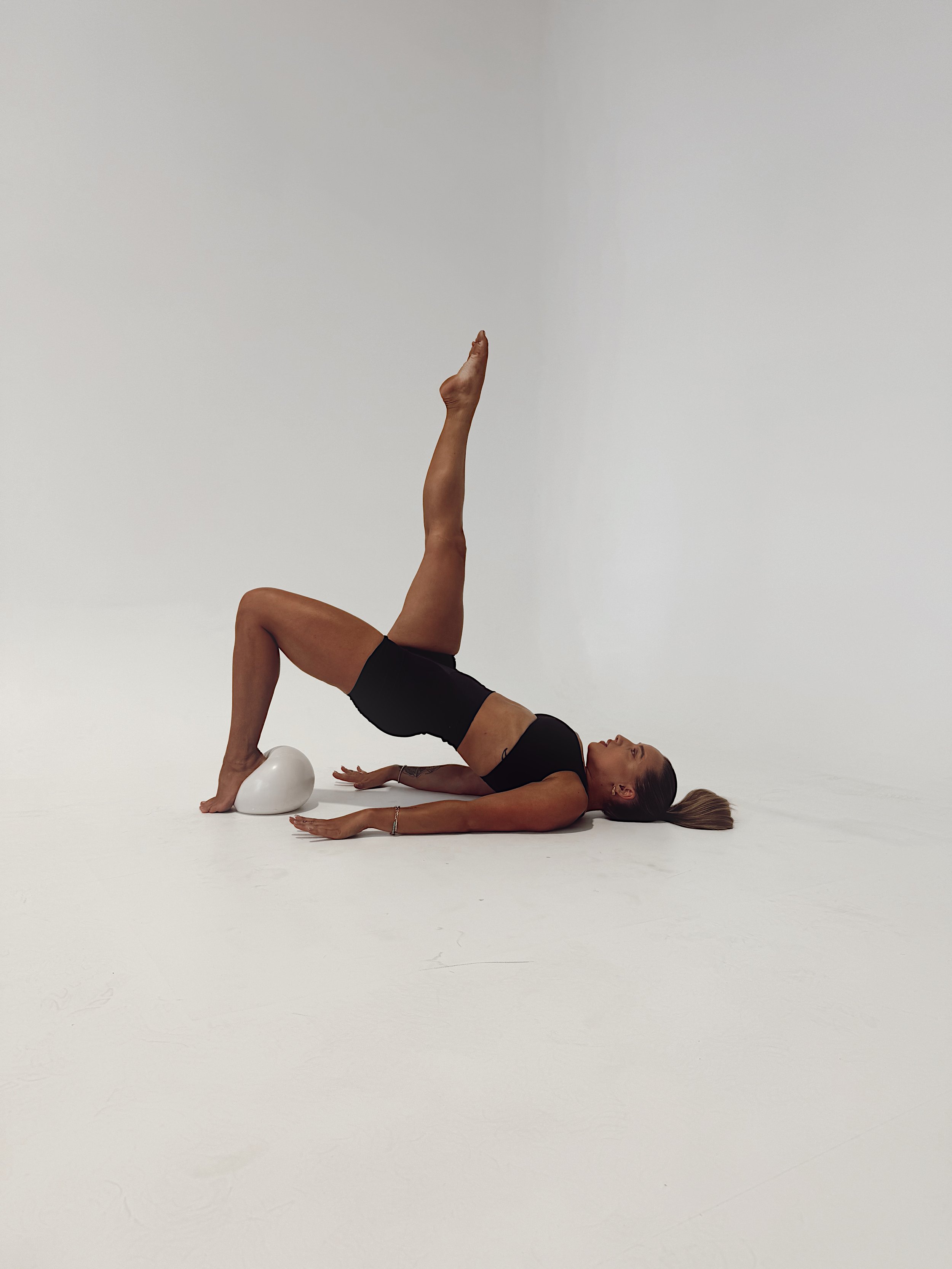 Person performing yoga bridge pose with one leg raised, on a white floor, using a yoga block for support.