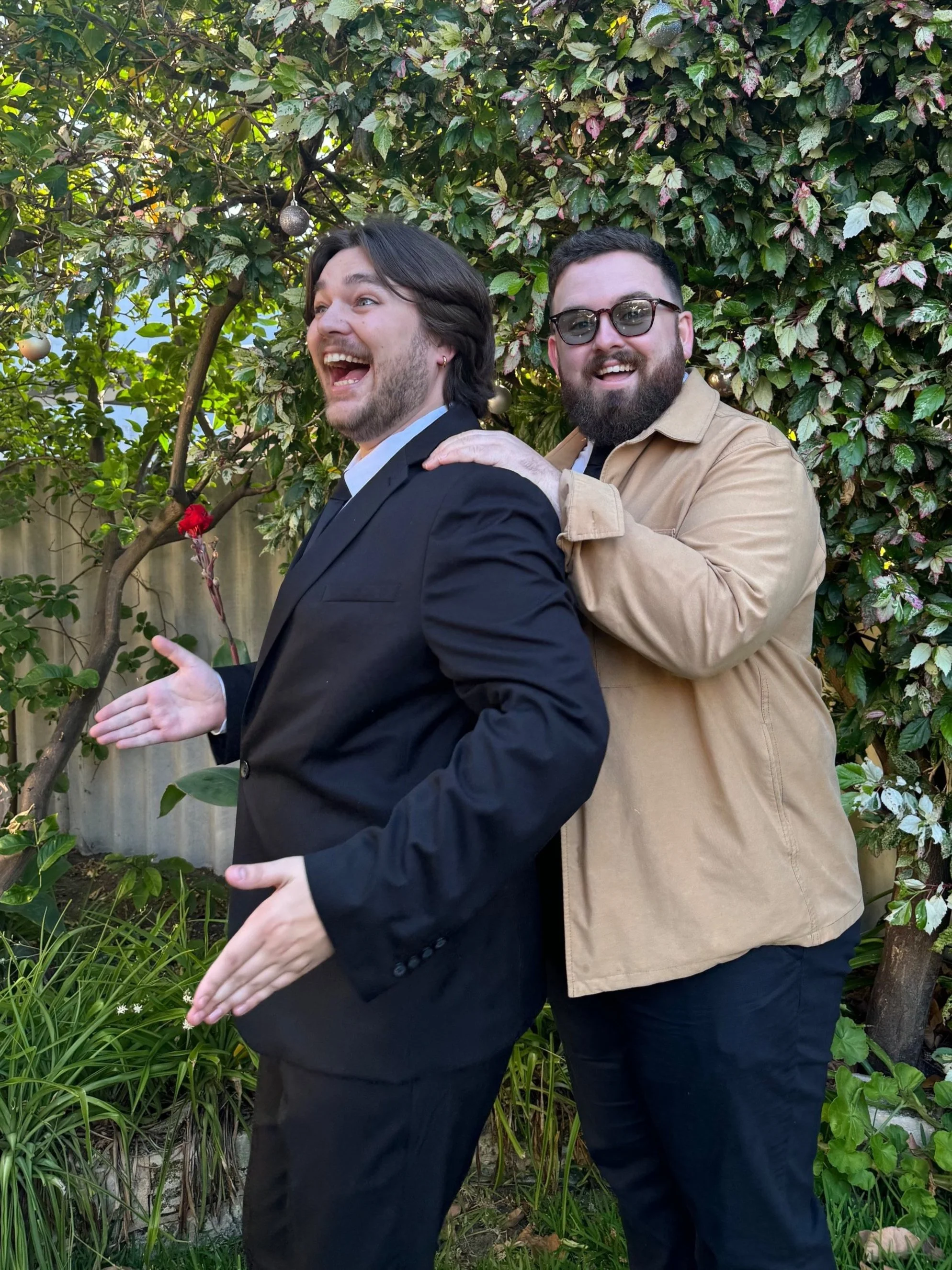 Two men posing playfully in a garden with lush black-green and variegated plants and trees. One man in a black suit is facing sideways with a surprised expression, gesturing with his hands. The other man, in a tan jacket and glasses, has a beard and is standing behind him, smiling and touching his shoulder.