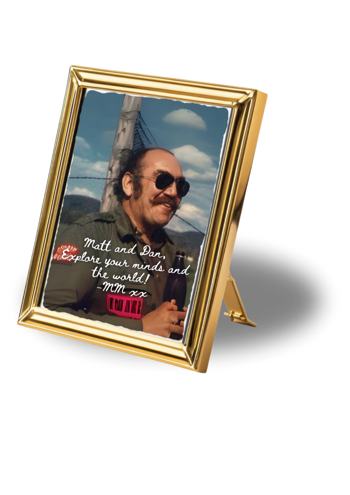 A framed photograph of a man with sunglasses, a beard, and a mustache, smiling outdoors against a cloudy sky and power lines. The photo has a handwritten message that says, "Matt and Dan, Explore your minds and the world! -MM xx".