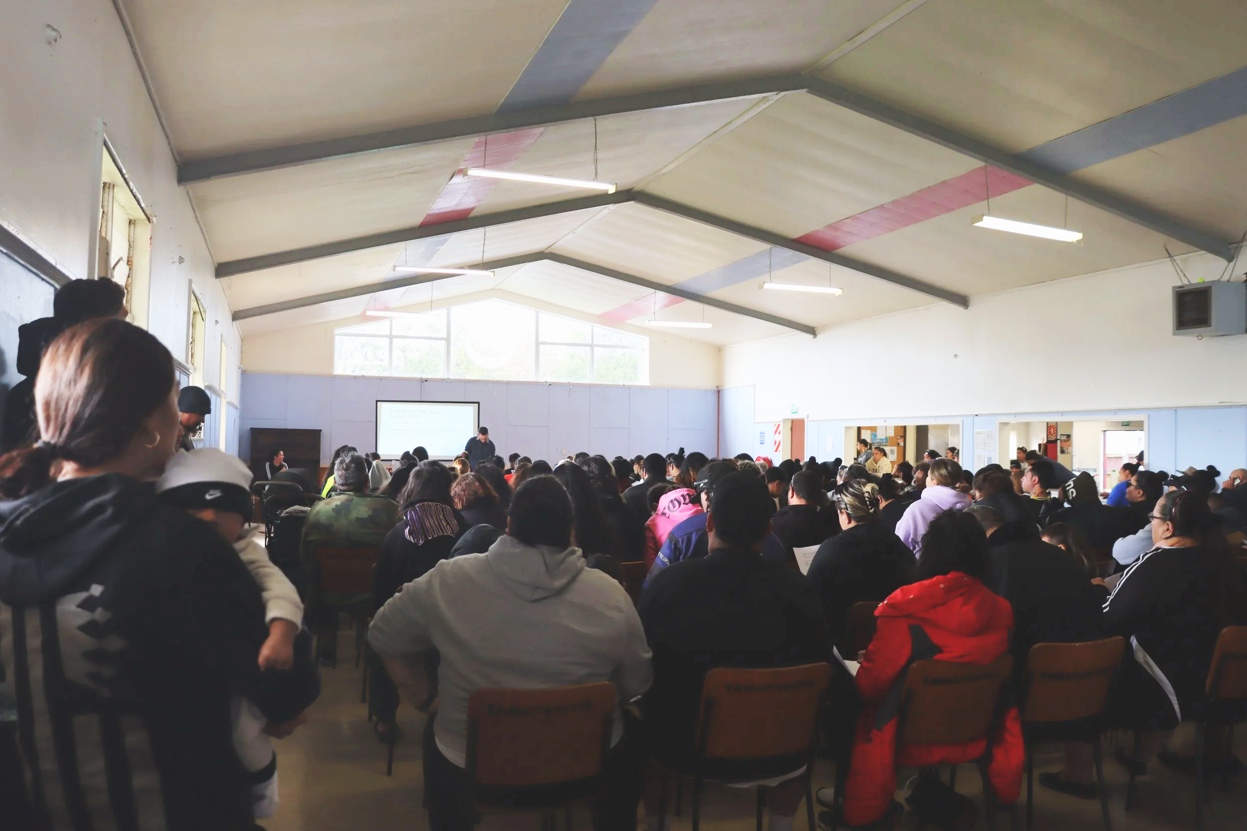 A large classroom or auditorium filled with students seated and listening to a presentation or lecture at the front. There is a projector screen and some windows letting in natural light.