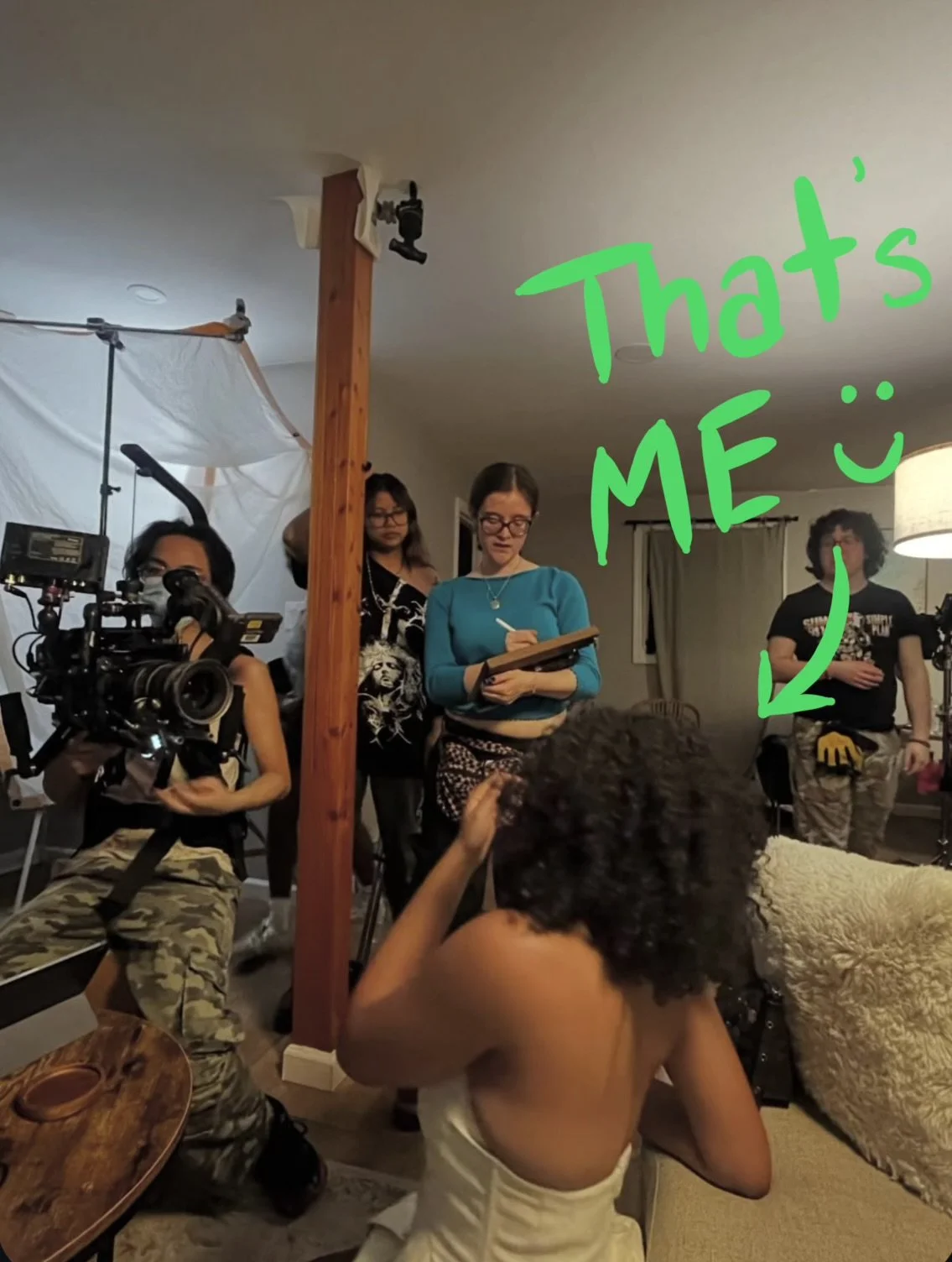 Behind the scenes of a film or photo shoot in a living room. Several crew members, including a woman with a camera, are present. A woman with glasses is taking notes, and another person is holding a walkie-talkie. The room is lit with professional lighting equipment. Green text with an arrow points to a man standing with yellow gloves and camouflage pants, with the words "That's ME :)" written.