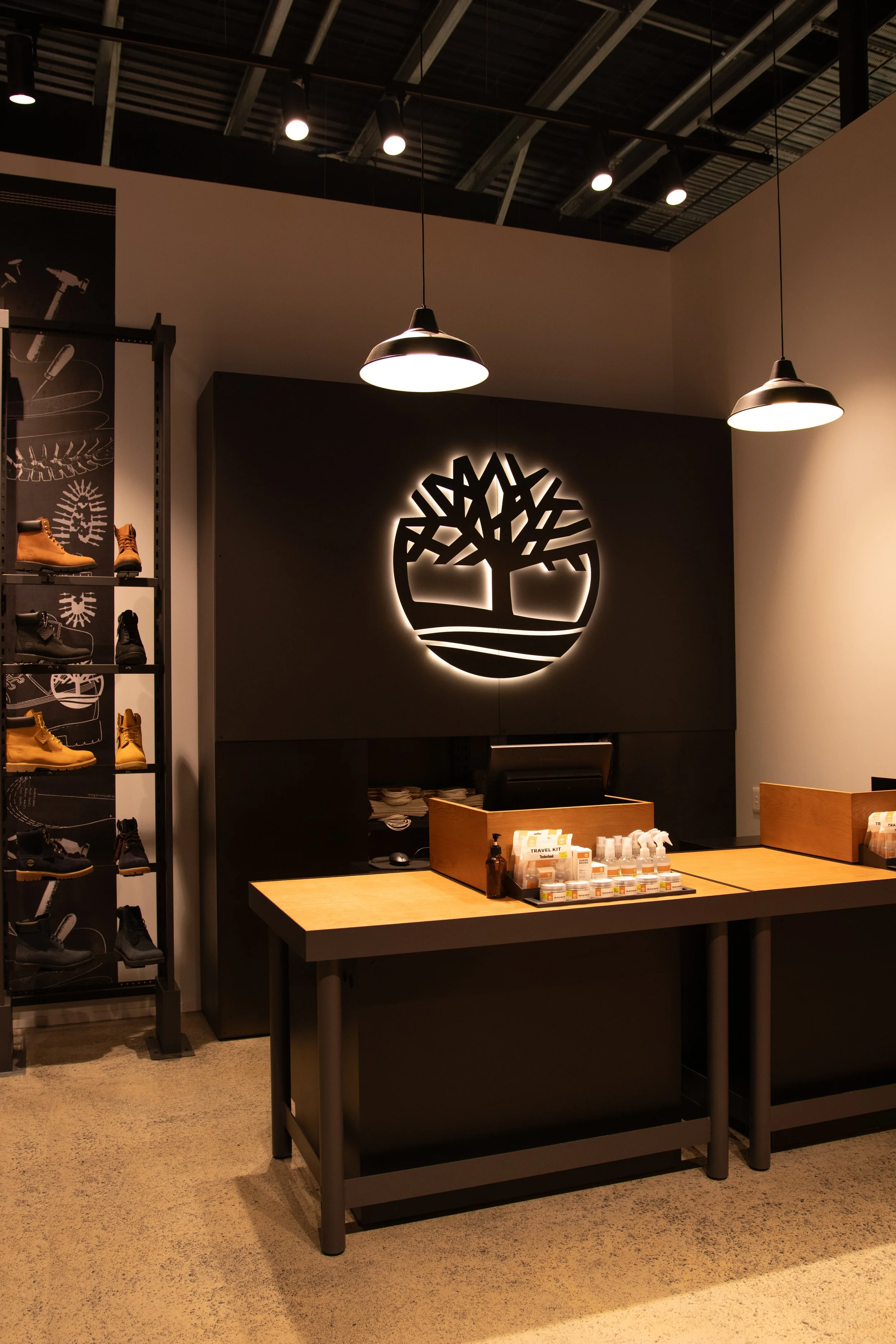 A shoe store interior with a large illuminated logo on the wall, shelves displaying boots, and a wooden counter with a register. Pendant lights hang from the ceiling.