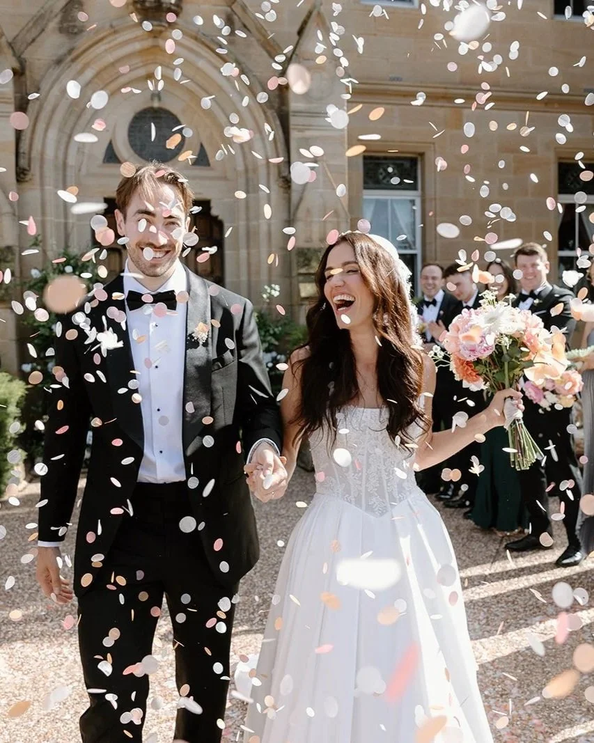 A bride and groom celebrating their wedding day with confetti falling around them outside a large stone building, with wedding guests in tuxedos in the background.