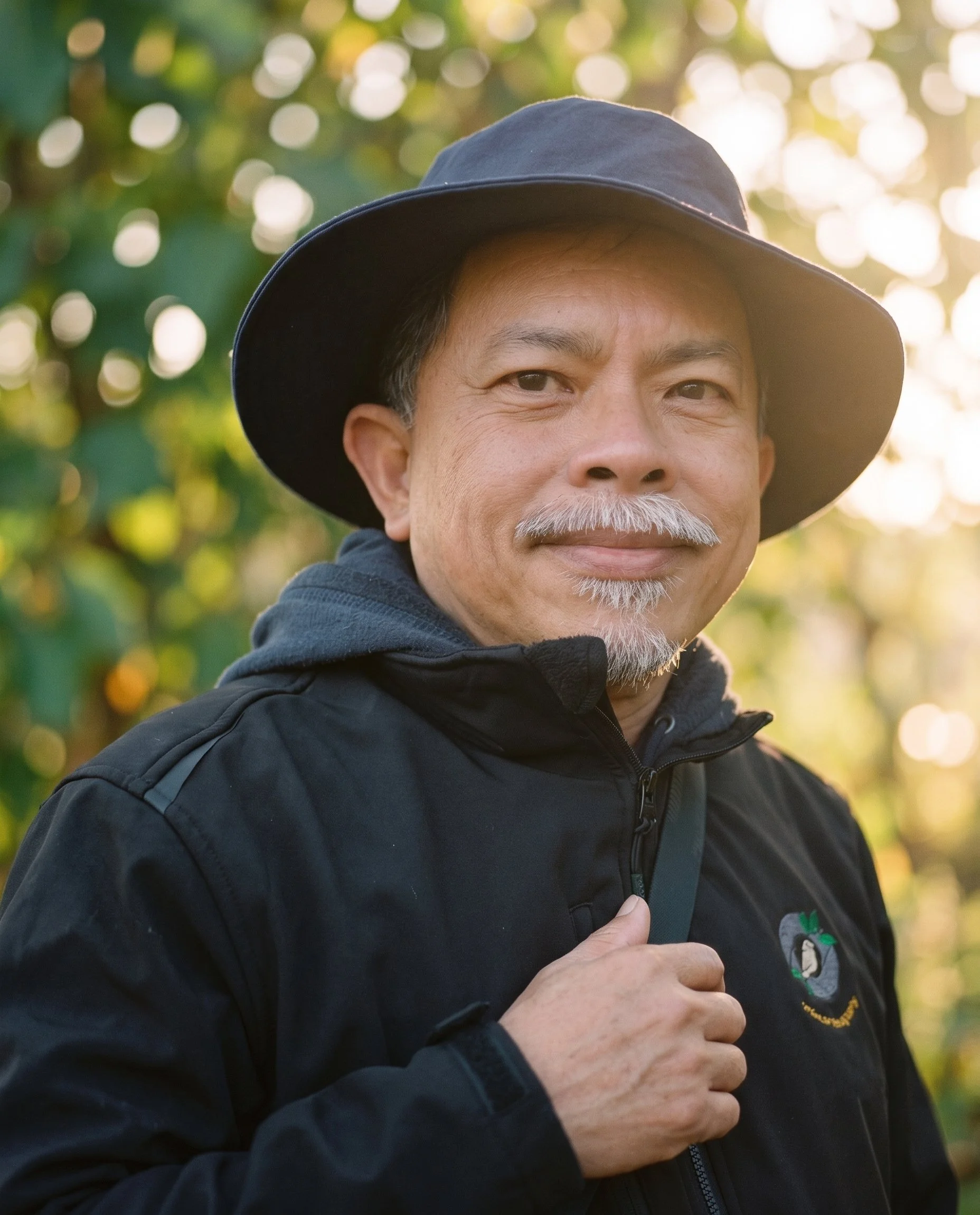 A portrait of an older man with a mustache and goatee, wearing a dark wide-brimmed hat and a black jacket with a small green and gray logo on the left chest, outdoors with a blurred background of sunlight filtering through trees.