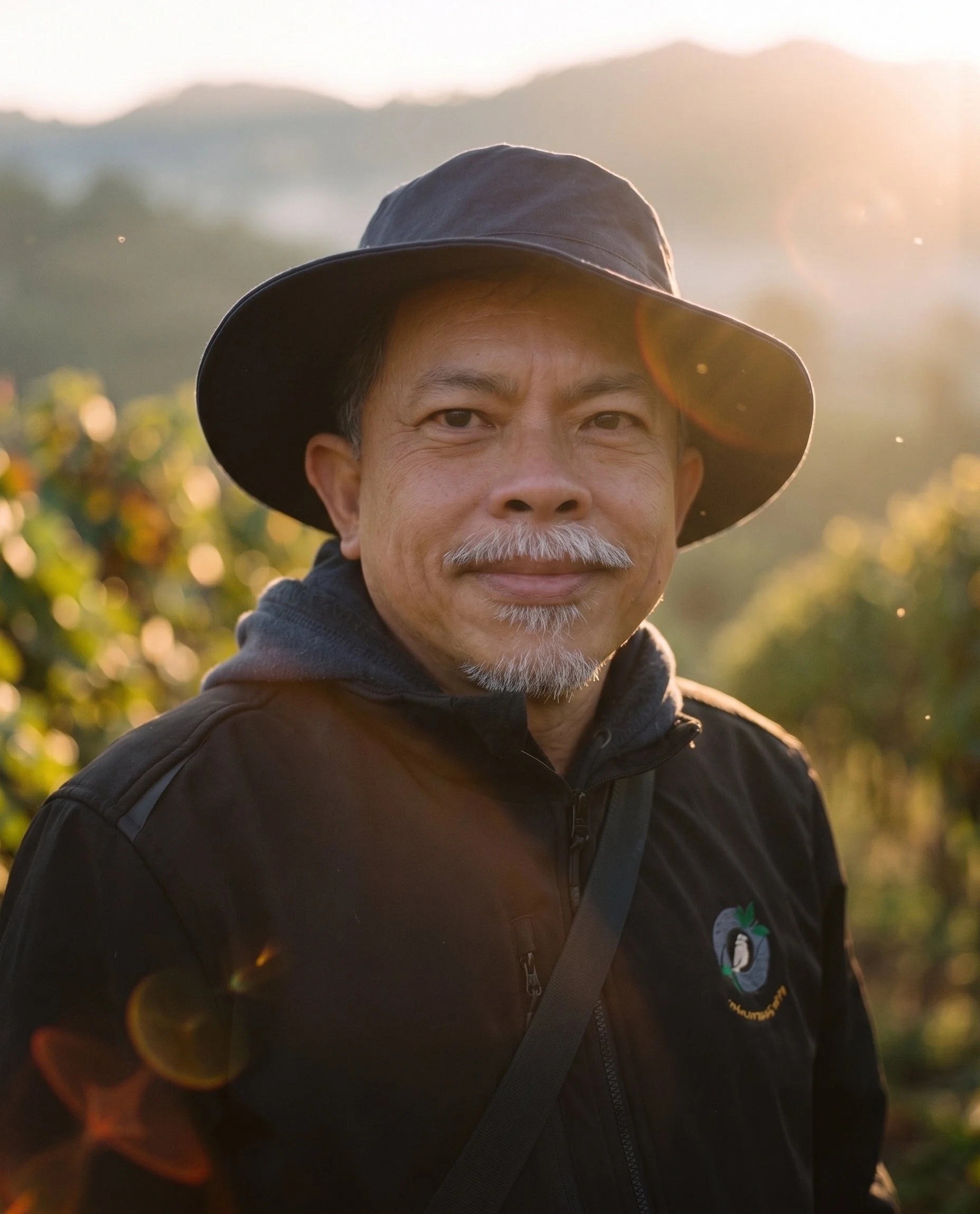 Close-up of an older man with gray hair and beard wearing a black wide-brimmed hat and black jacket, outdoors during sunset.