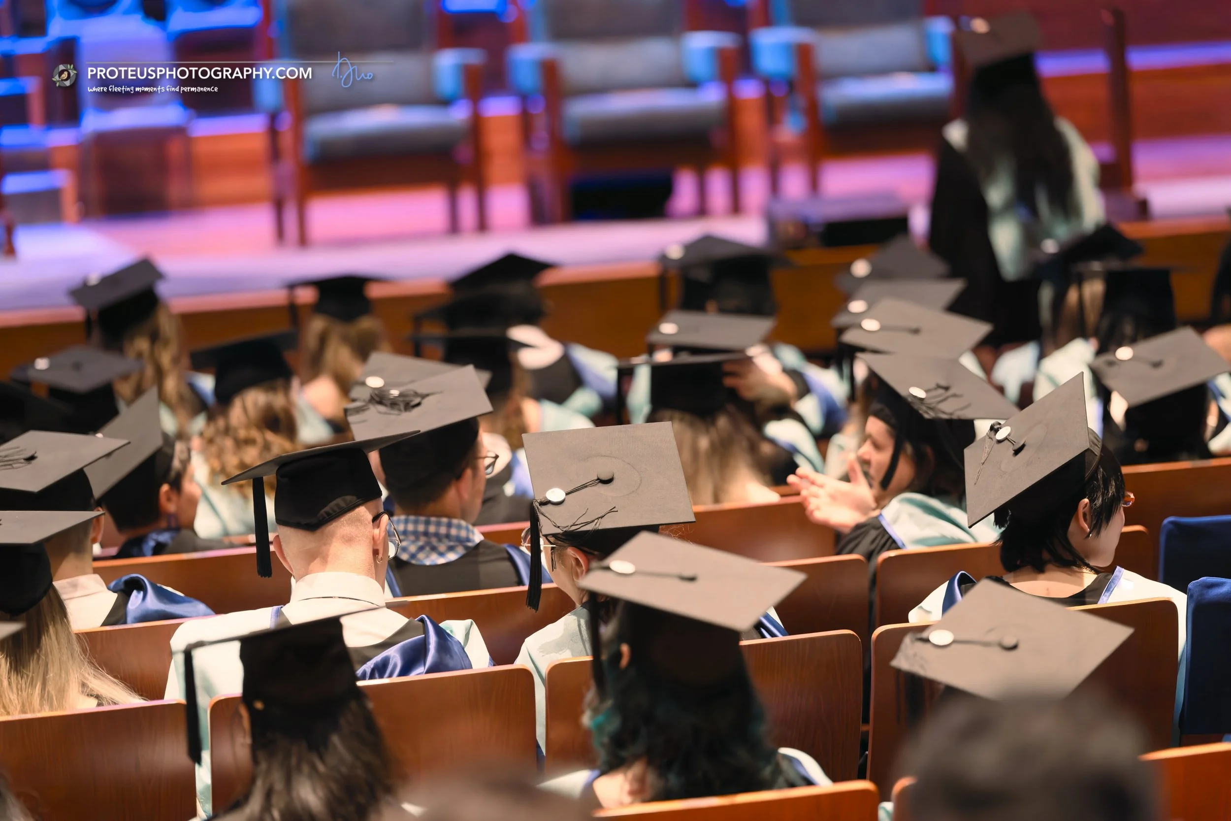 College graduation ceremony with students wearing black caps and gowns, seated in an auditorium.