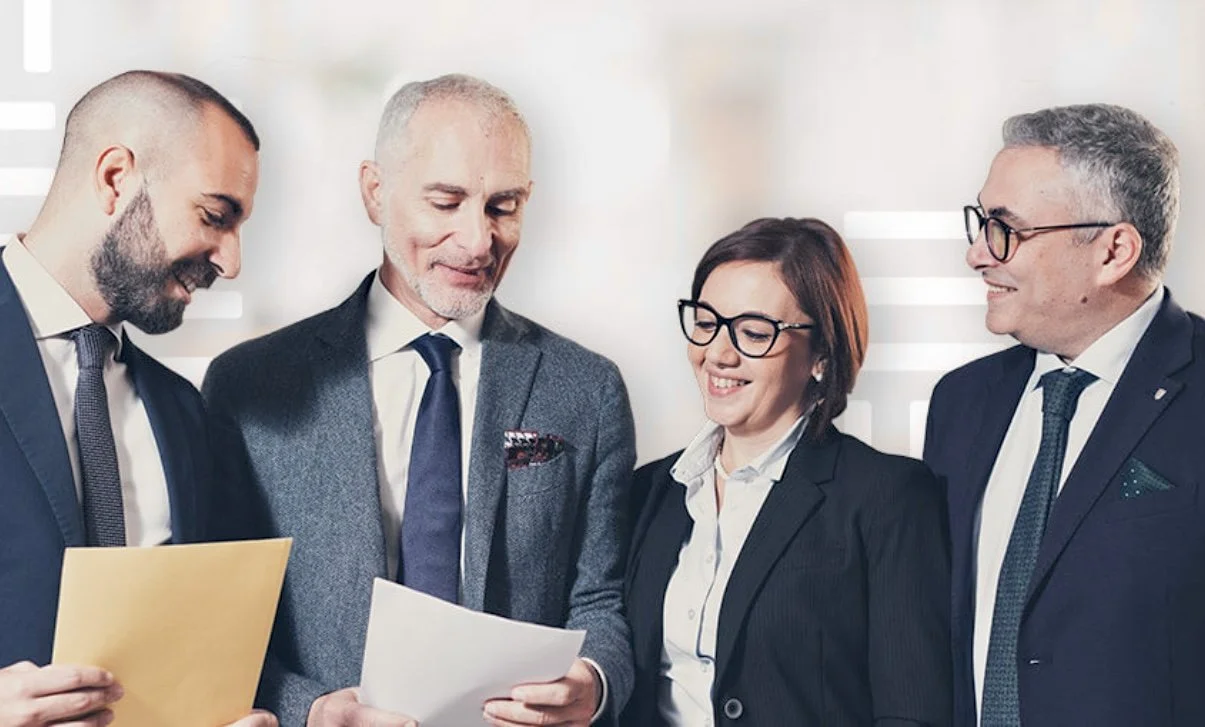 Quattro professionisti sorridenti in abiti formali, che si confrontano su documenti in un ufficio luminoso.