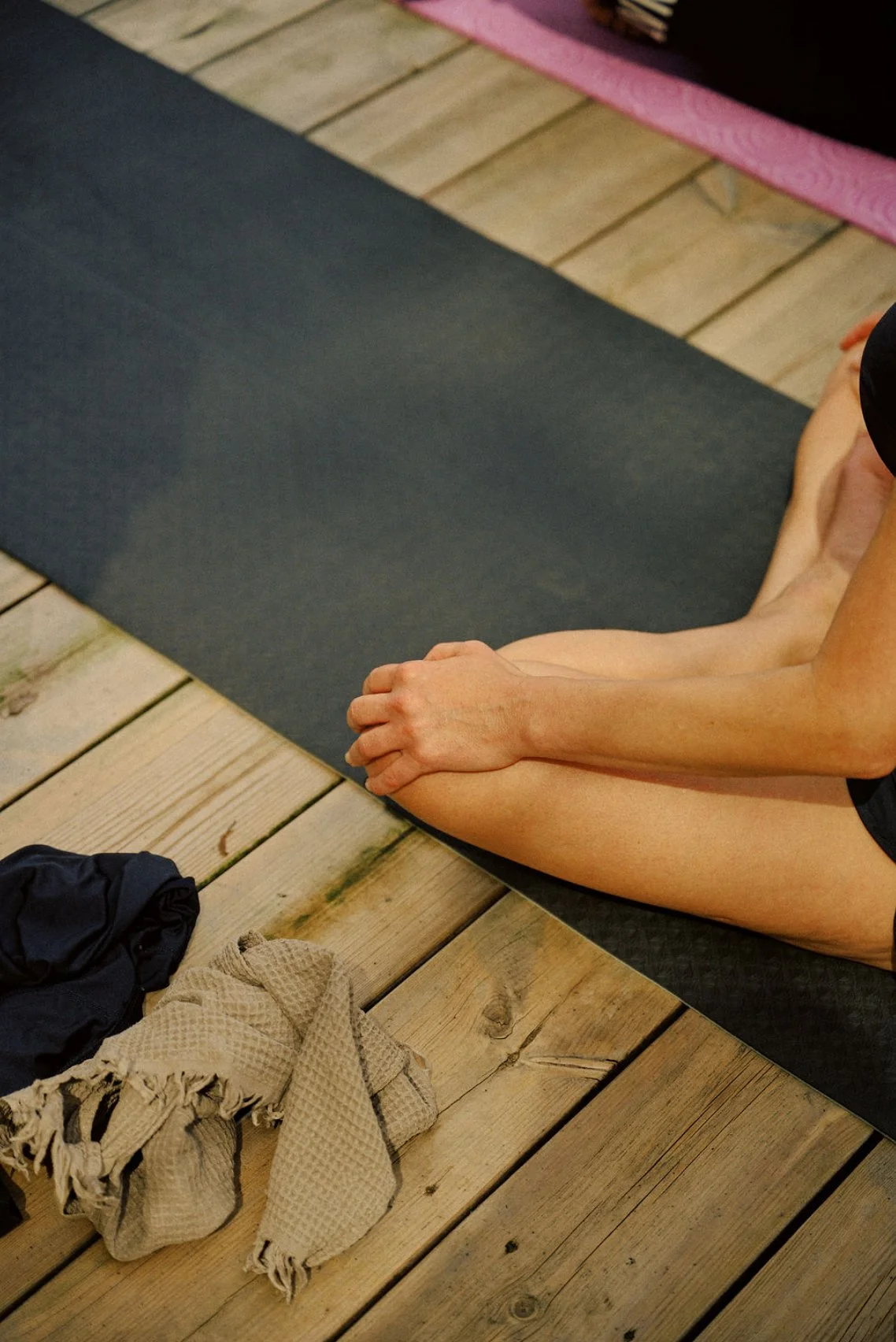 Person sitting on a yoga mat on a wooden deck, preparing for yoga or meditation. Clothing and other items are on the deck nearby.