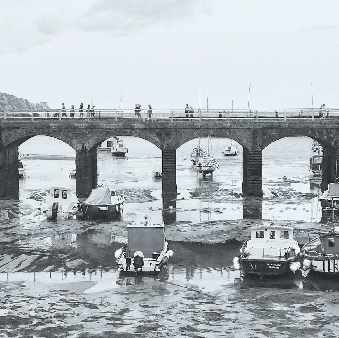 People walking along a pier with boats docked below, in a black and white photo.