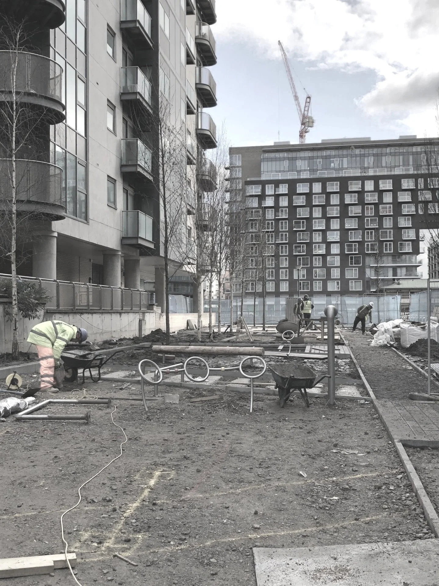 Construction workers working on a site in an urban area with tall apartment buildings in the background, some trees, construction equipment, and materials scattered around.