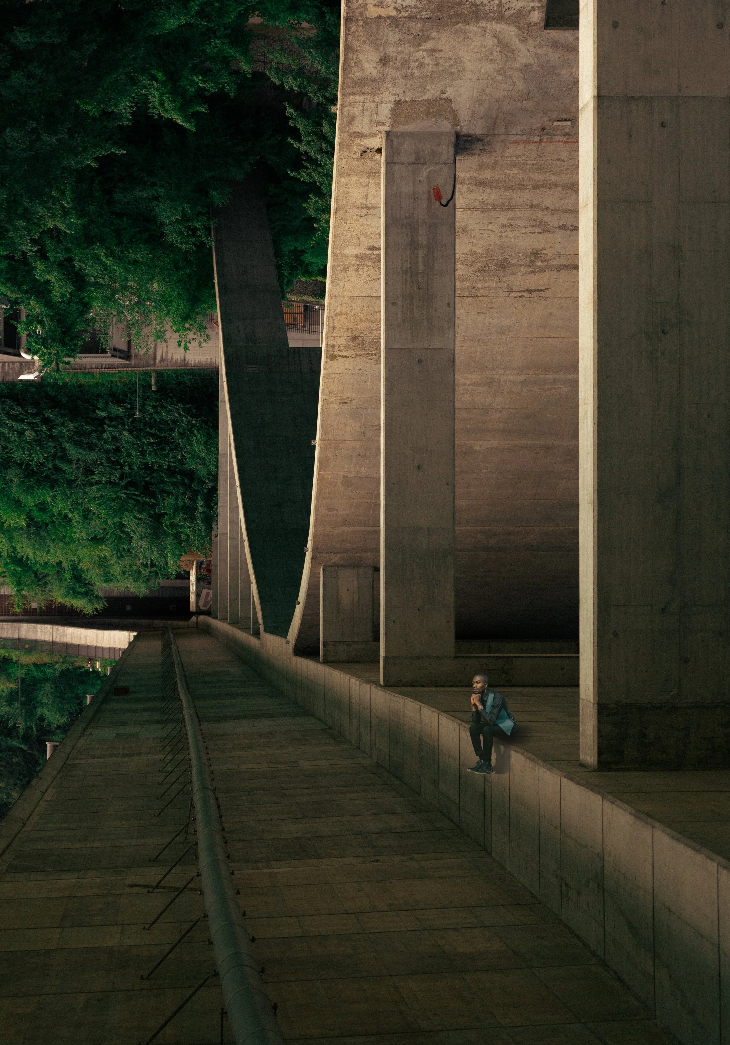 Ein Mann sitzt einsam auf einer hohen, unzugänglichen Betonmauer in einer modernen, urbanen Umgebung mit grünen Bäumen im Hintergrund, bei Nacht.