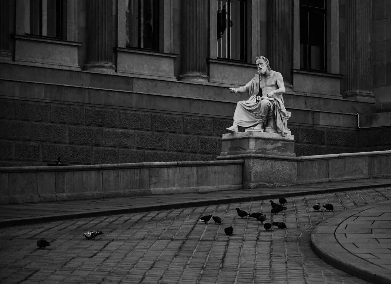 Schwarz-Weiß-Foto einer Statue eines sitzenden Mannes vor einem historischen Gebäude mit Säulen. Mehrere Tauben sitzen auf dem gepflasterten Gehweg im Vordergrund.