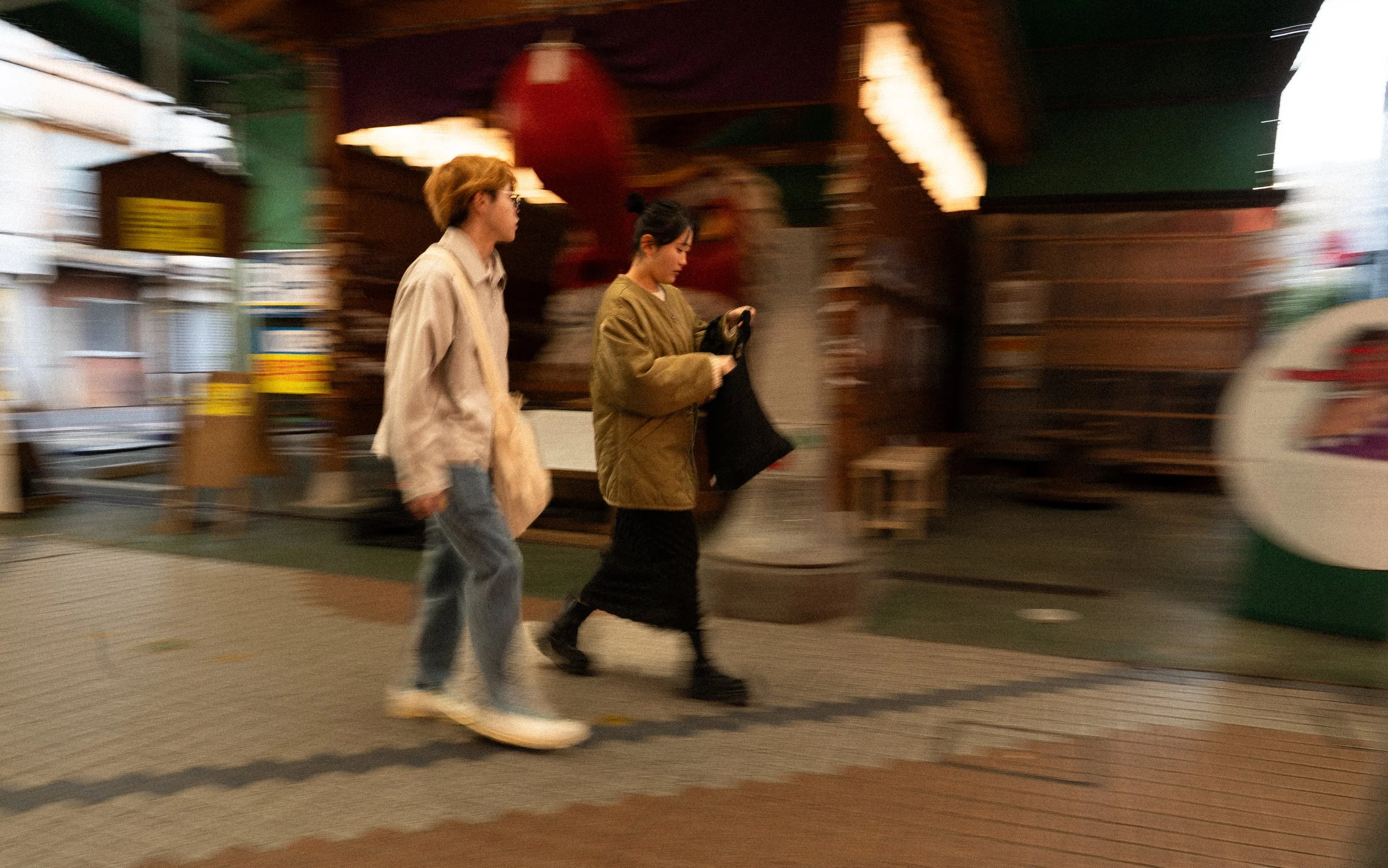 Zwei junge Frauen gehen nebeneinander an einem überdachten, farbenfrohen Marktstand vorbei.