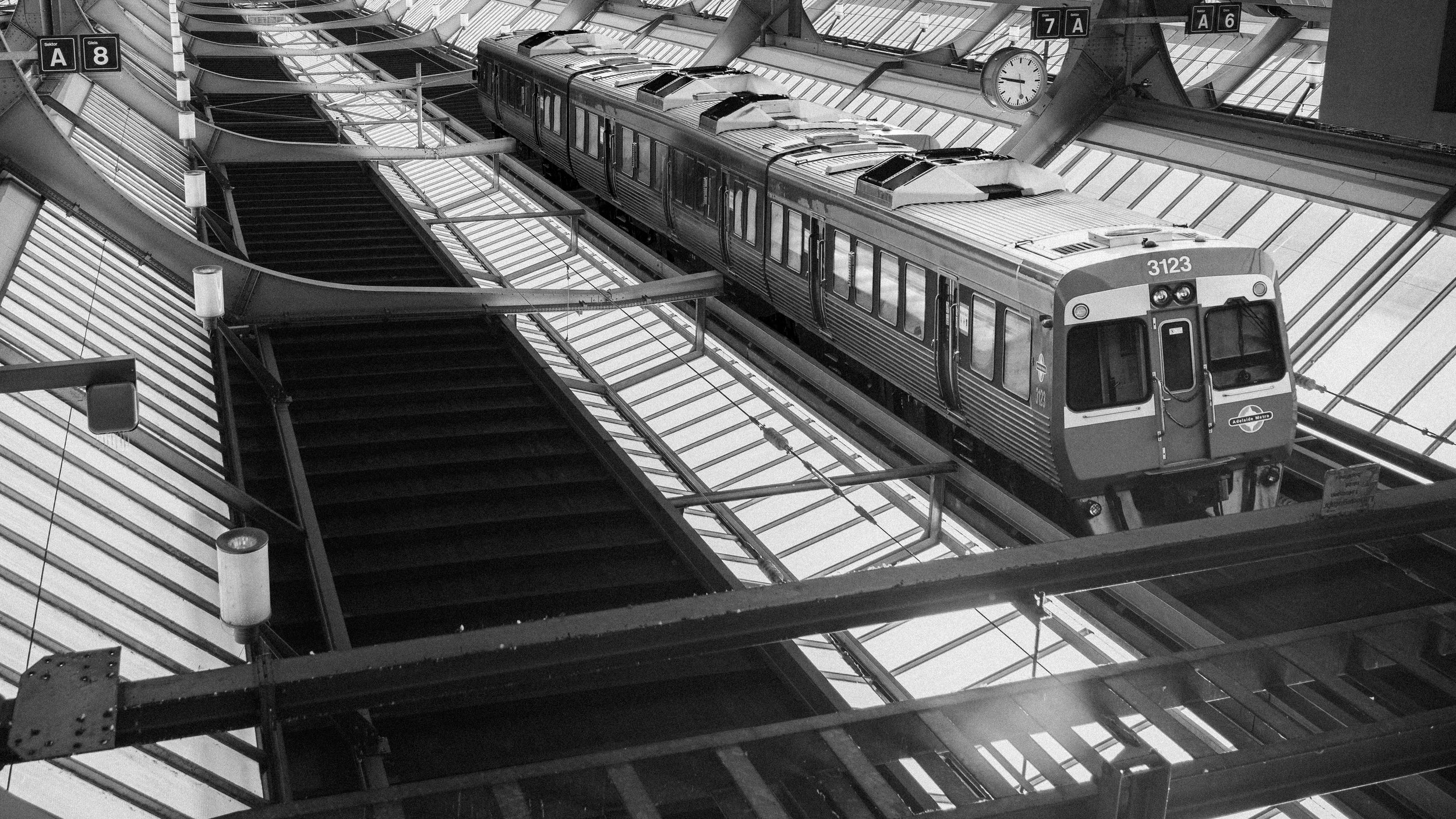 Ein grauer Zugfahrzeug hält auf einer erhöhten Plattform in einem modernen Bahnhof, umgeben von Glasstrukturen und Treppen, mit analogen Uhren und Informationsschildern im Hintergrund.