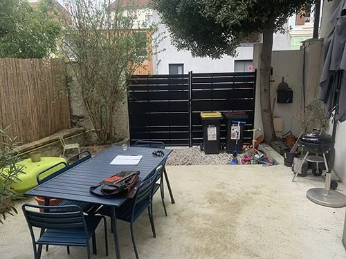 Un jardin avec une table en métal noir, plusieurs chaises, un barbecue, une clôture en bois et un mur avec un mur noir en arrière-plan.