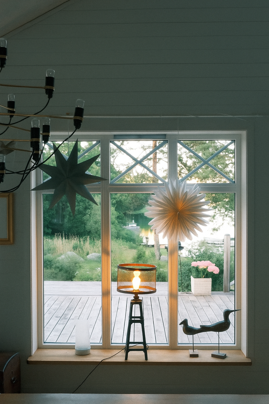 Interior view of a windowed room with decorative paper stars hanging from the ceiling, a small table lamp, a bottle-shaped diffuser, a ceramic bird sculpture, and a view of a pond and trees outside.