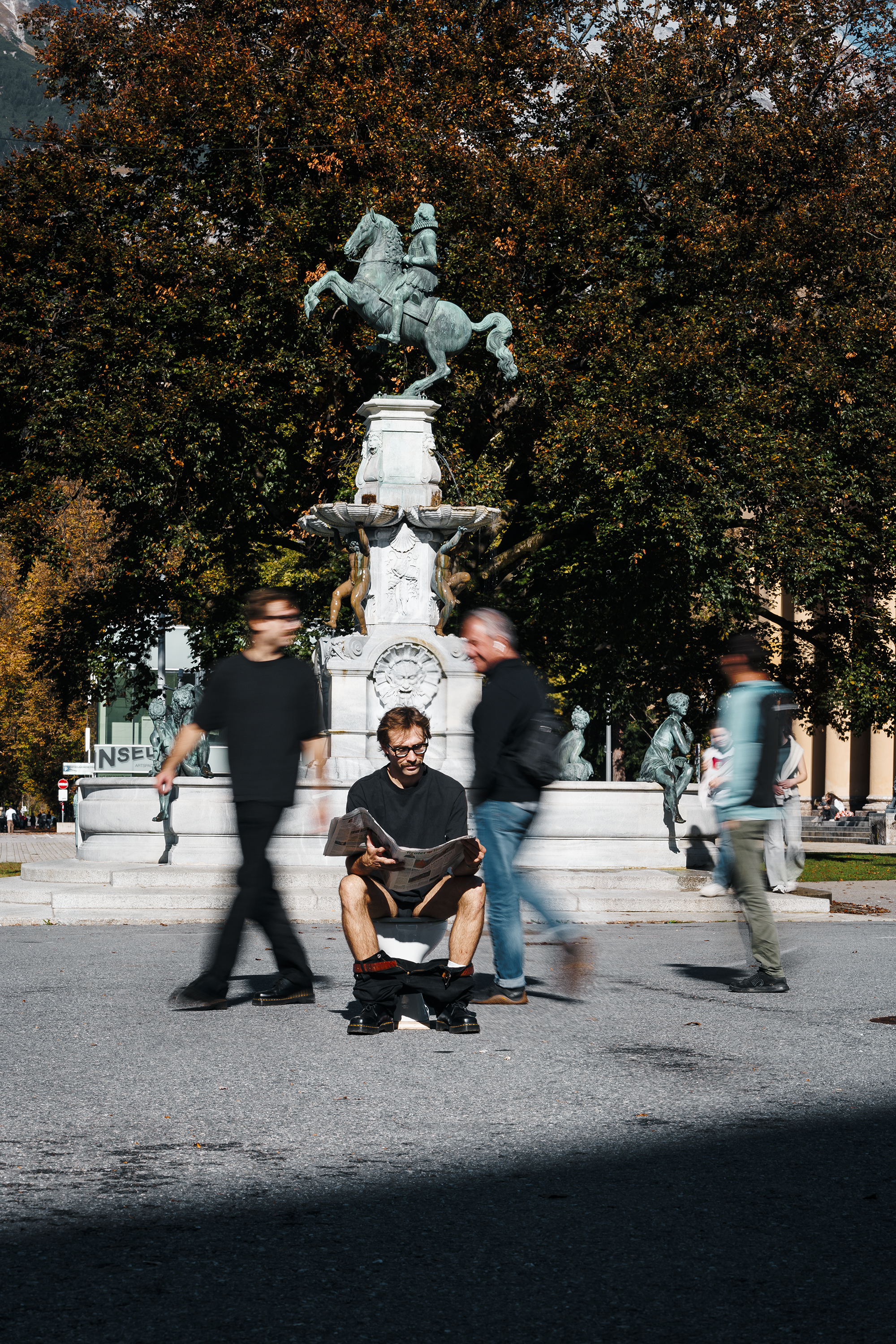Ein Mann sitzt auf einer Bank und liest eine Zeitung vor einer Brunnenstatue mit einem Reiter. Mehrere Personen gehen an ihm vorbei, während sich im Hintergrund Bäume und weitere Statuen befinden.