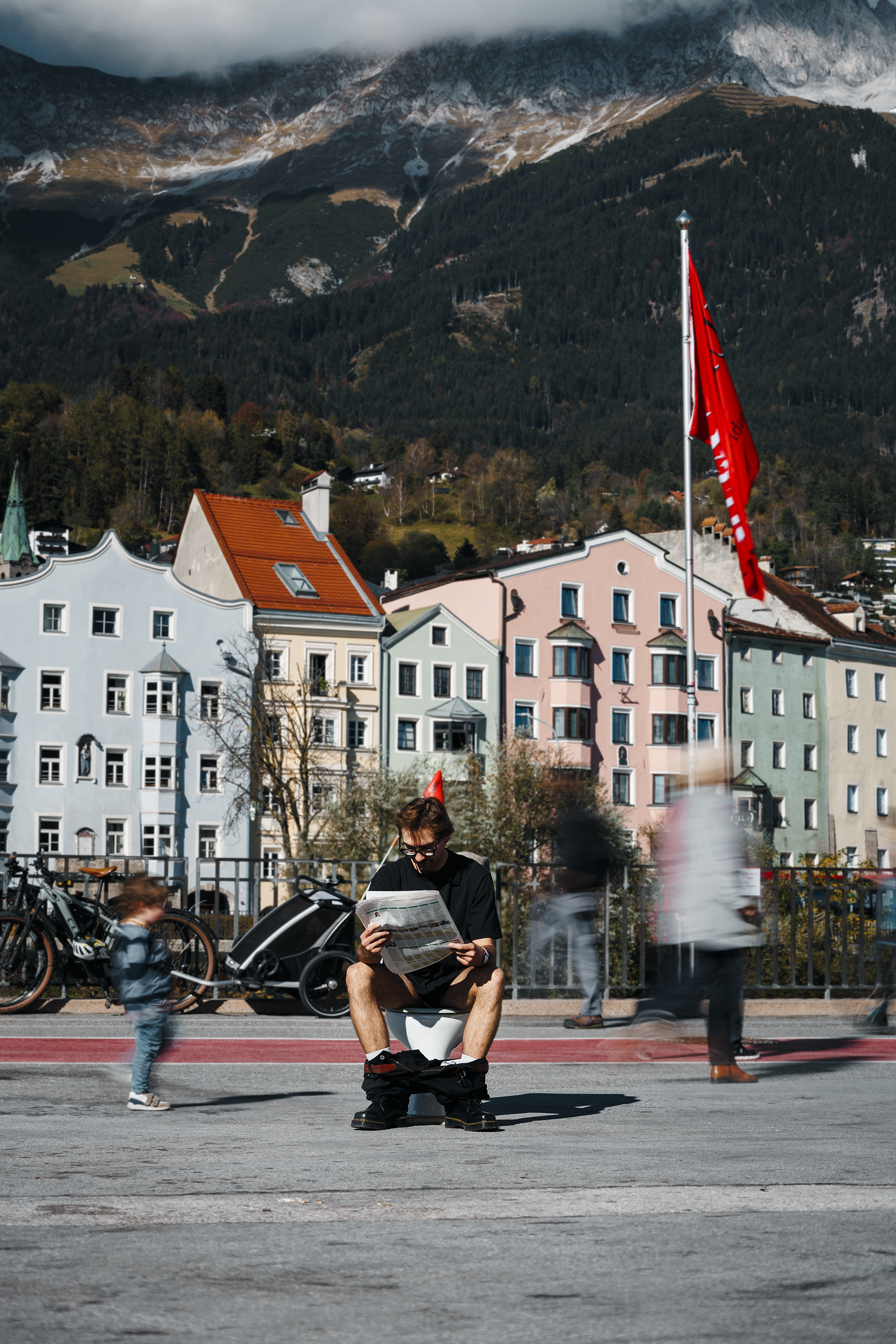 Junger Mann sitzt auf einer Toilette und liest eine Zeitung, während er auf einer Straße in einer Stadt mit bunten Gebäuden im Hintergrund sitzt. Im Vordergrund sind laufende Passanten und ein kleines Kind zu sehen.