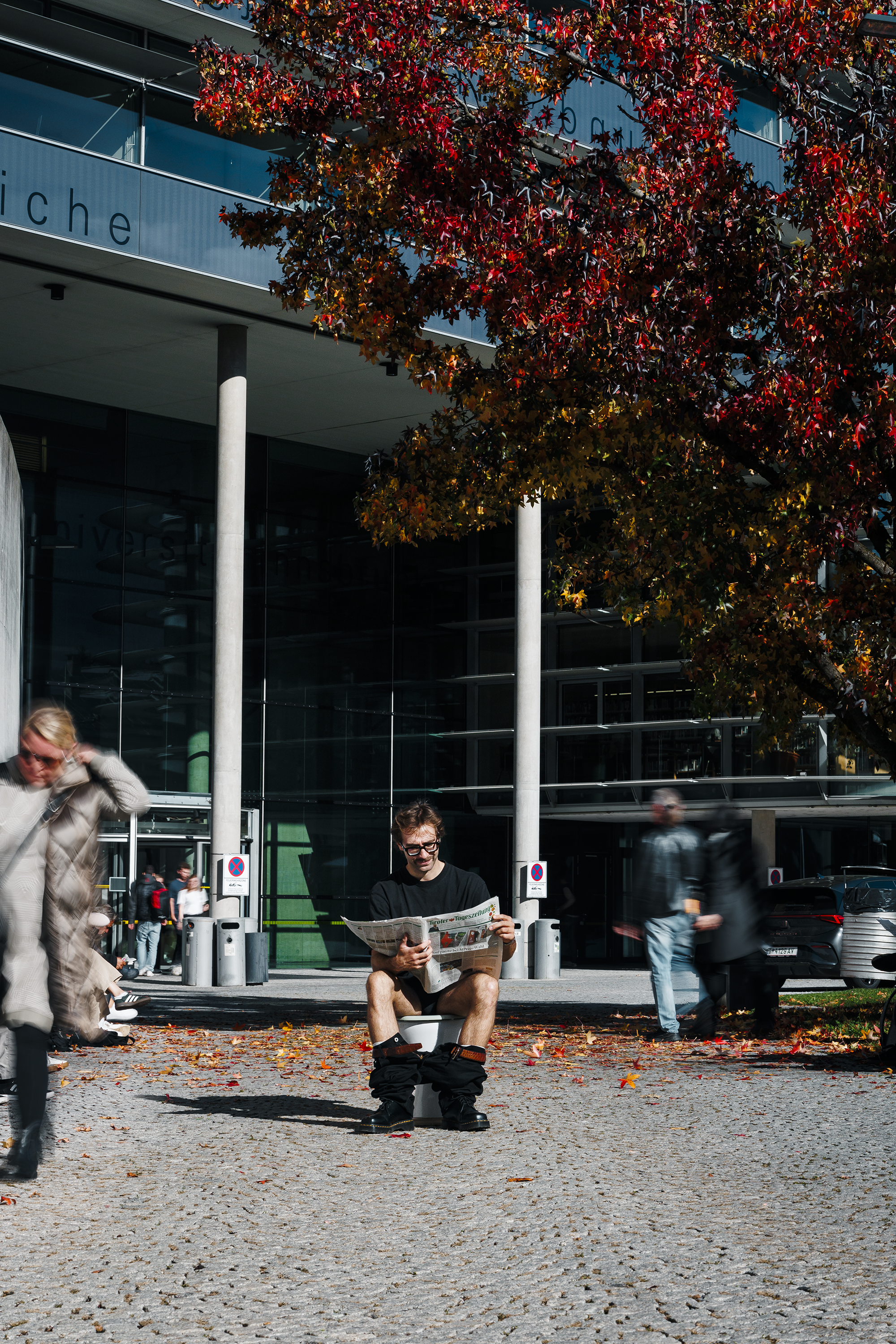 Ein junger Mann sitzt im Freien auf einer Toilette und liest eine Zeitung. Es ist Herbst, die Bäume sind rot und gelb gefärbt. Mehrere Menschen gehen vorbei, einige sind unscharf und bewegen sich schnell.
