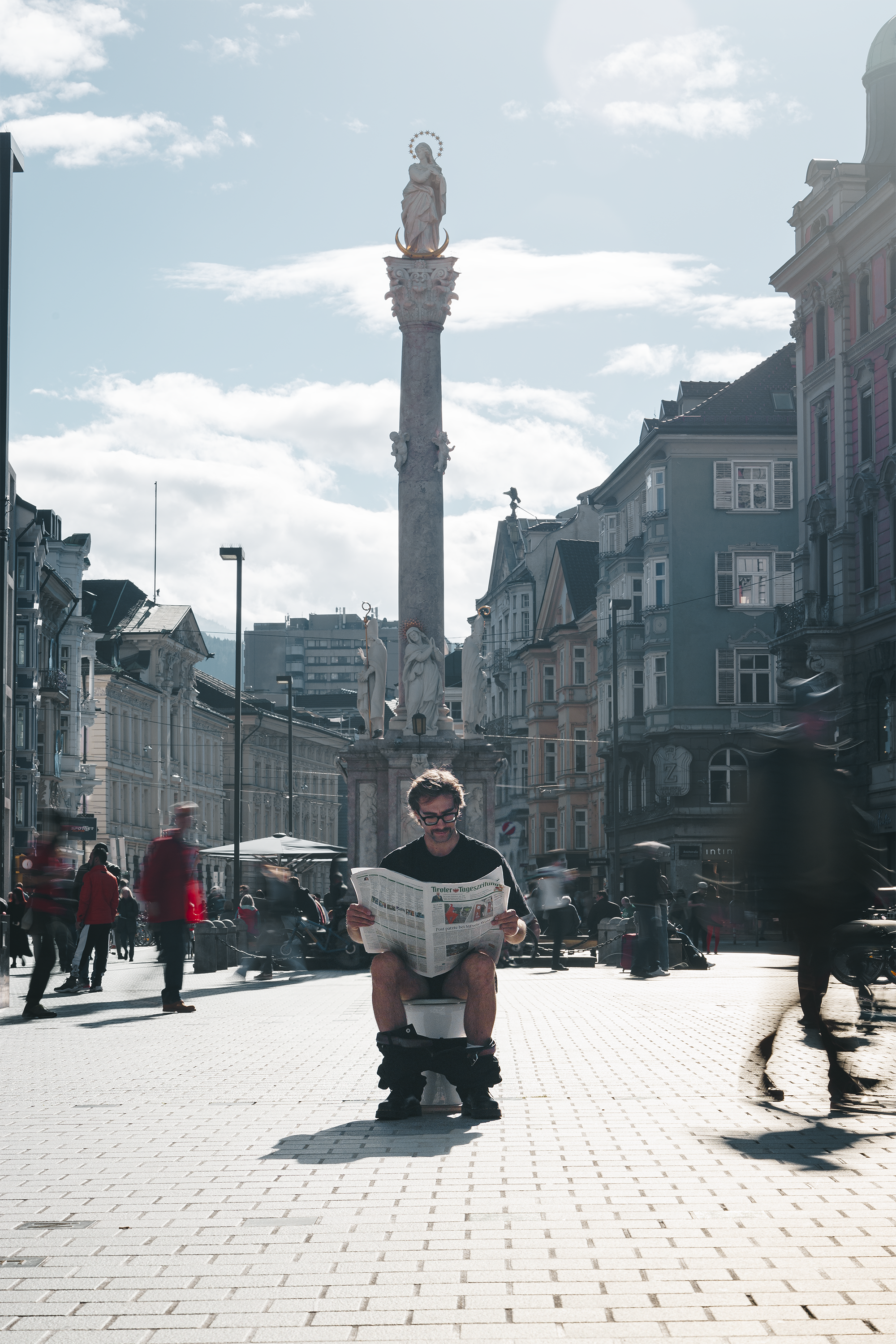 Ein Mann sitzt auf einer Toilette in einer belebten Stadtstraße und liest eine Zeitung. Im Hintergrund ist eine barocke Statue auf einem hohen Säulenmonument zu sehen, umgeben von historischen Gebäuden und Menschen, die vorbeigehen.