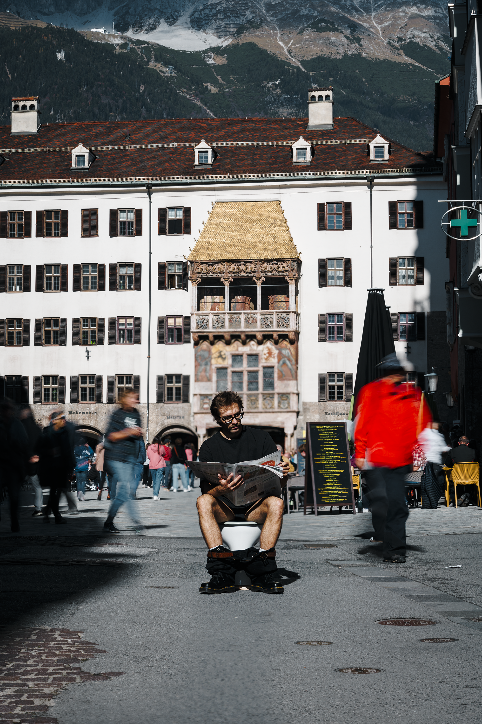 Ein junger Mann sitzt auf einem Klositz in der Stadt und liest eine Zeitung. Im Hintergrund sind Menschen, die durch einen Platz laufen, sowie ein altes, mehrstöckiges Gebäude mit einem barocken Balkon und einem roten Ziegeldach. Es herrscht lebhafte Atmosphäre, mit einigen Menschen, die draußen sitzen, und einem bewölkten Himmel mit Berg im Hintergrund.