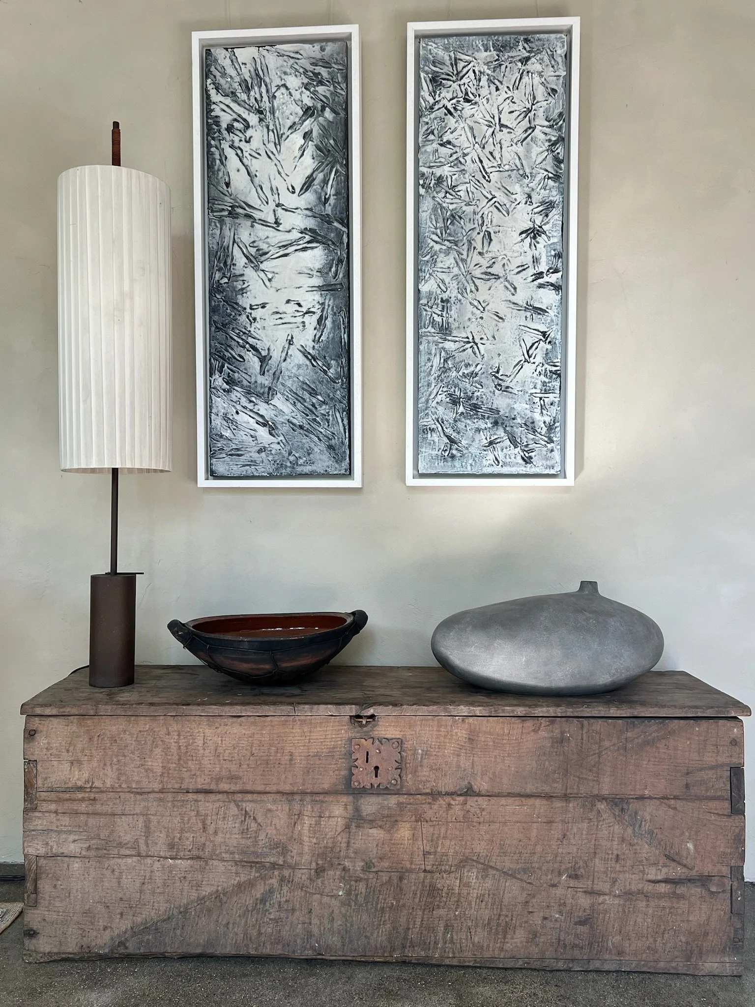 Ein rustikaler Holzkastentisch mit einer schwarzen und einer grauen Skulptur, daneben eine Lampe und zwei vertikale abstrakte Kunstwerke mit schwarzen, weißen und grauen Mustern an der Wand.