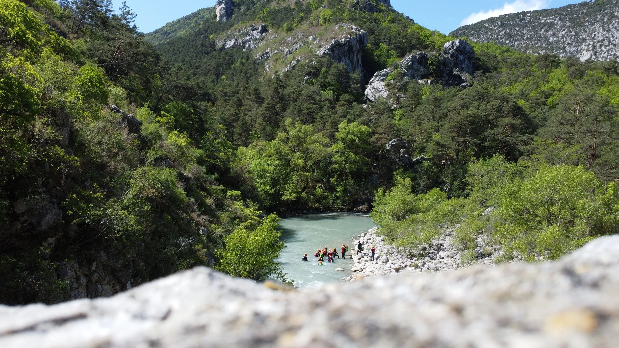 Eine Gruppe von Menschen, die in einem Fluss in einer grünen, bergigen Landschaft wandern oder schwimmen, umgeben von hohen Bergen und dichten Binen.
