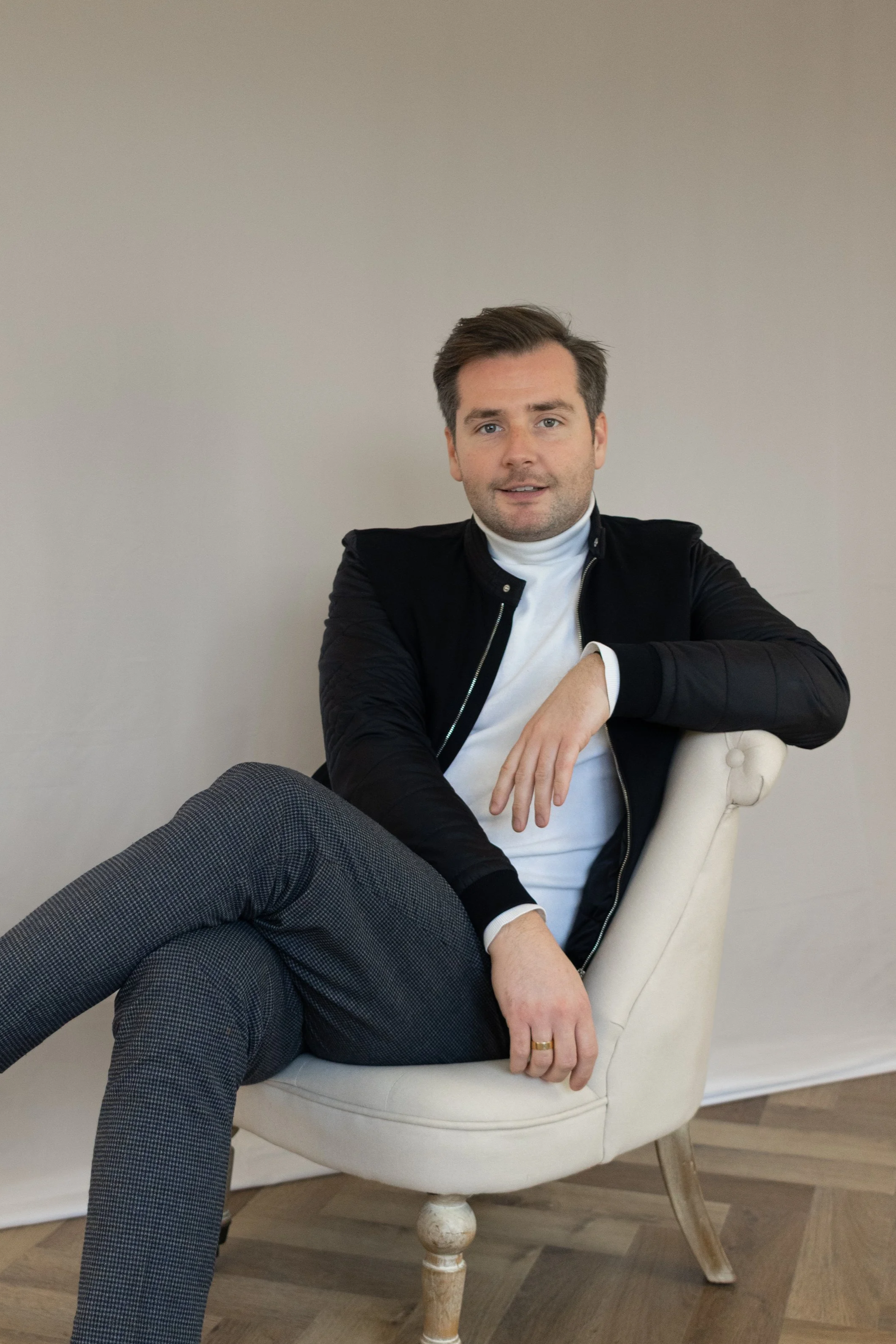 Ein Mann sitzt entspannt in einem weißen Sessel vor einer hellen Wand, trägt ein weißes T-Shirt, eine schwarze Jacke und graue Hosen, hat braune Haare und schaut in die Kamera.