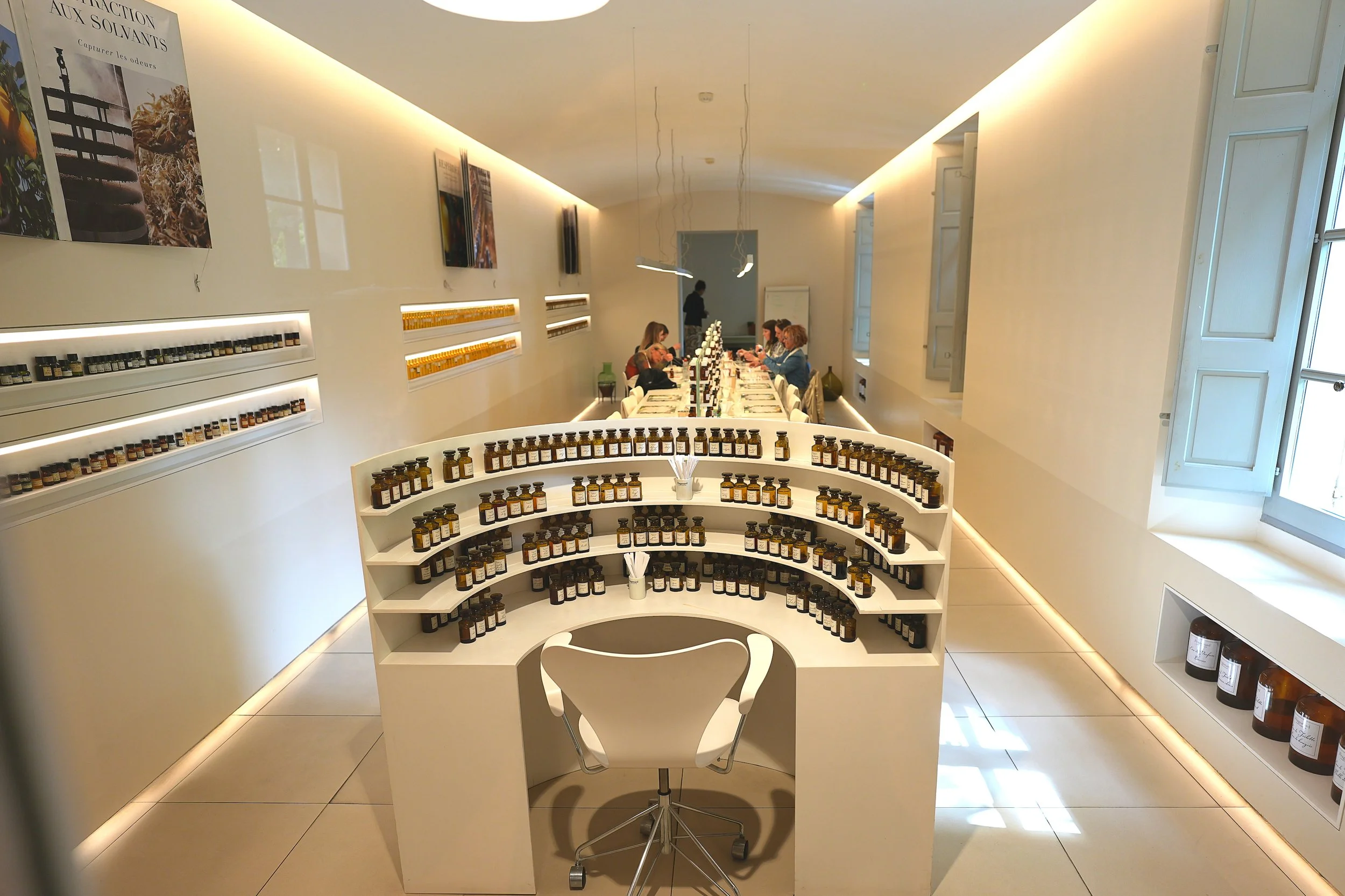 Innenraum eines Raums mit weißen Regalen und einem langen Tisch, an dem mehrere Menschen sitzen, sowie zahlreiche kleine Flaschen und Gläser auf den Regalen