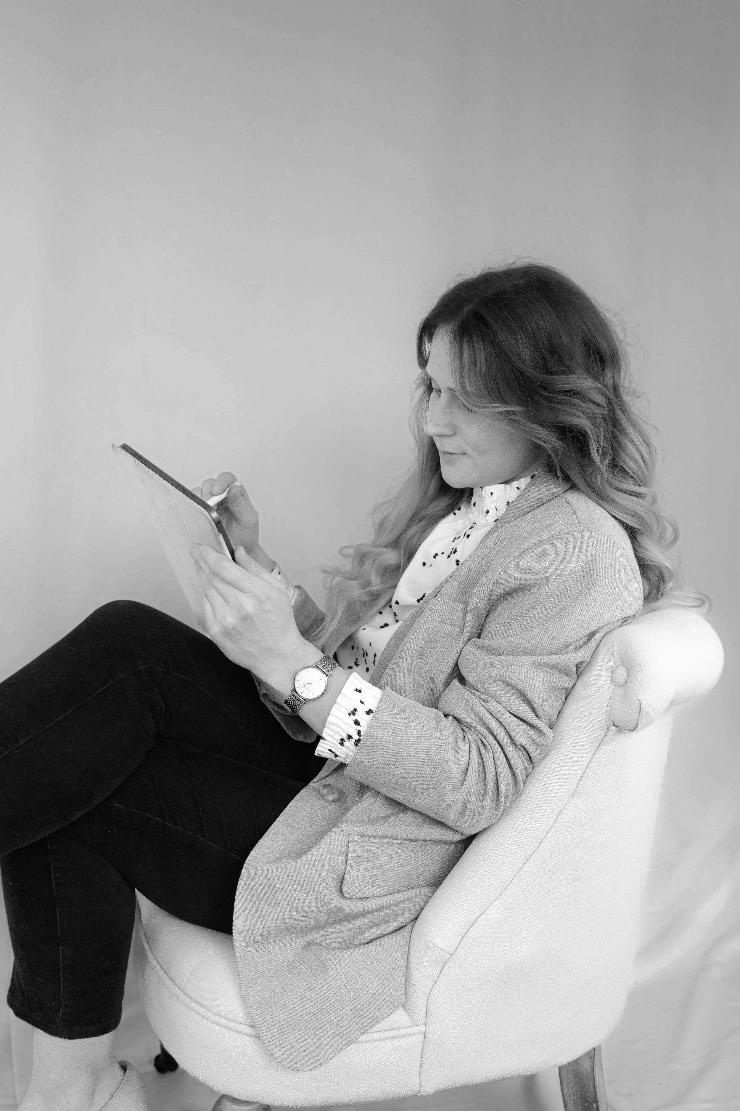 Eine Frau mit langem, lockigem Haar sitzt seitlich in einem Stuhl und liest ein Tablet. Sie trägt eine Armbanduhr und einen hellgrauen Blazer, ein weißes Hemd mit schwarzen Punkten und schwarze Hosen.