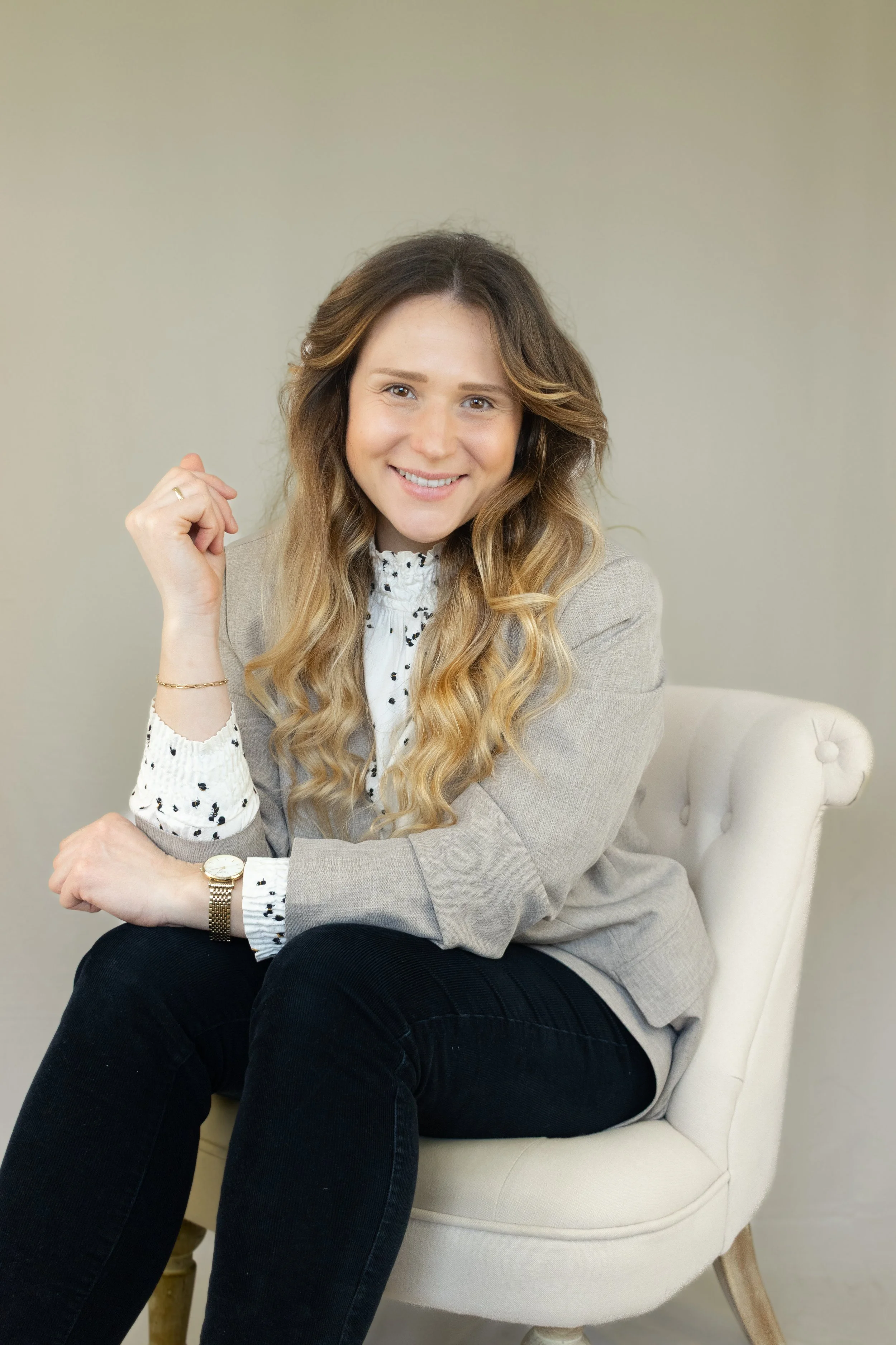 Lächelnde Frau mit langen, gewellten Haaren in einem grauen Blazer und weißem Hemd, sitzend auf einem weißen Stuhl.