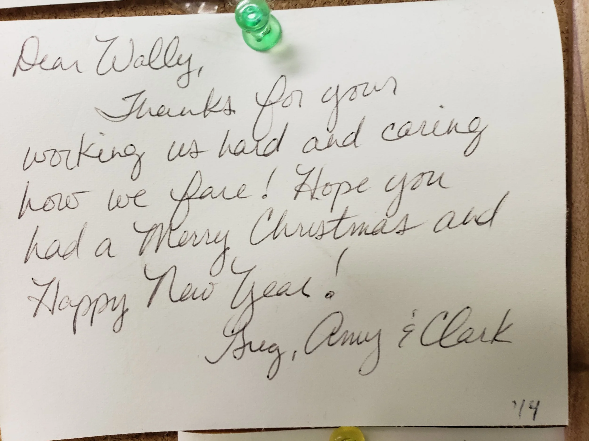 Handwritten Christmas and New Year greeting note pinned with a green pushpin, thanking Wally for his work and wishing him Merry Christmas and a Happy New Year, signed by Greg, Amy, and Clark.