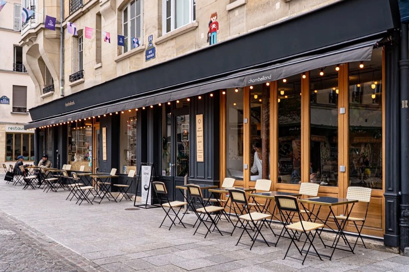 Une terrasse de restaurant avec des tables et des chaises en plein air, située devant un bâtiment avec des fenêtres en bois et des guirlandes lumineuses, dans une rue pavée.