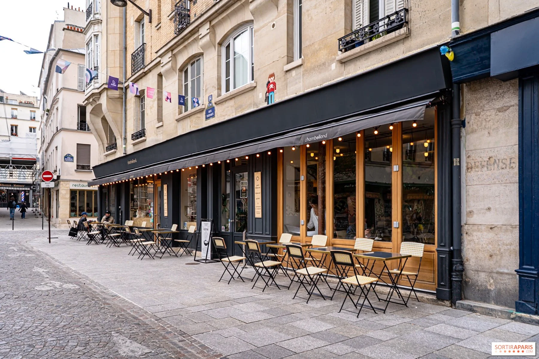 Terrasse de café avec tables et chaises devant un bâtiment en pierre avec une devanture noire et des fenêtres en bois, à Paris.